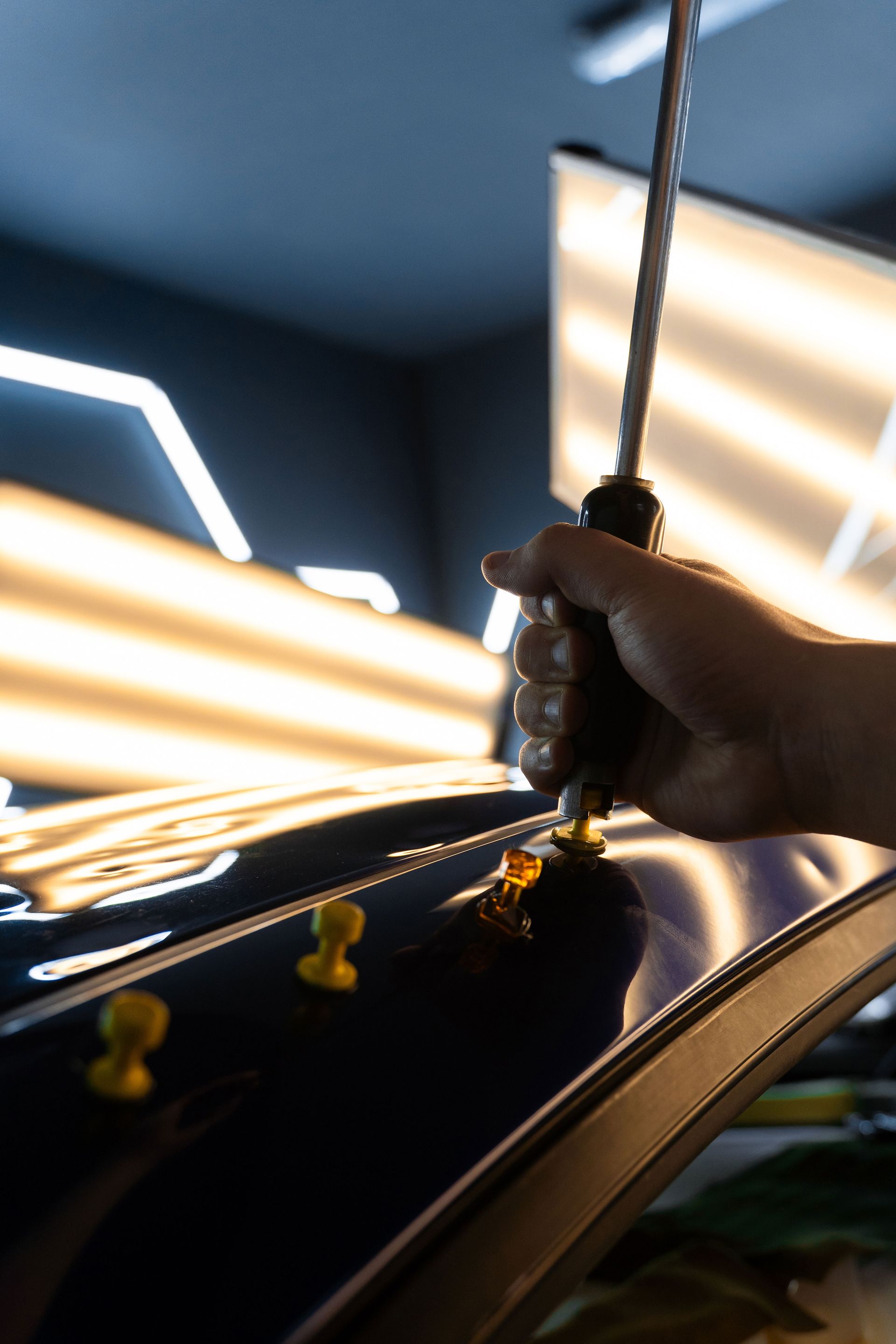 A technician uses a tool to pull a dent from a dark blue car body in a workshop with specialized strip lighting.