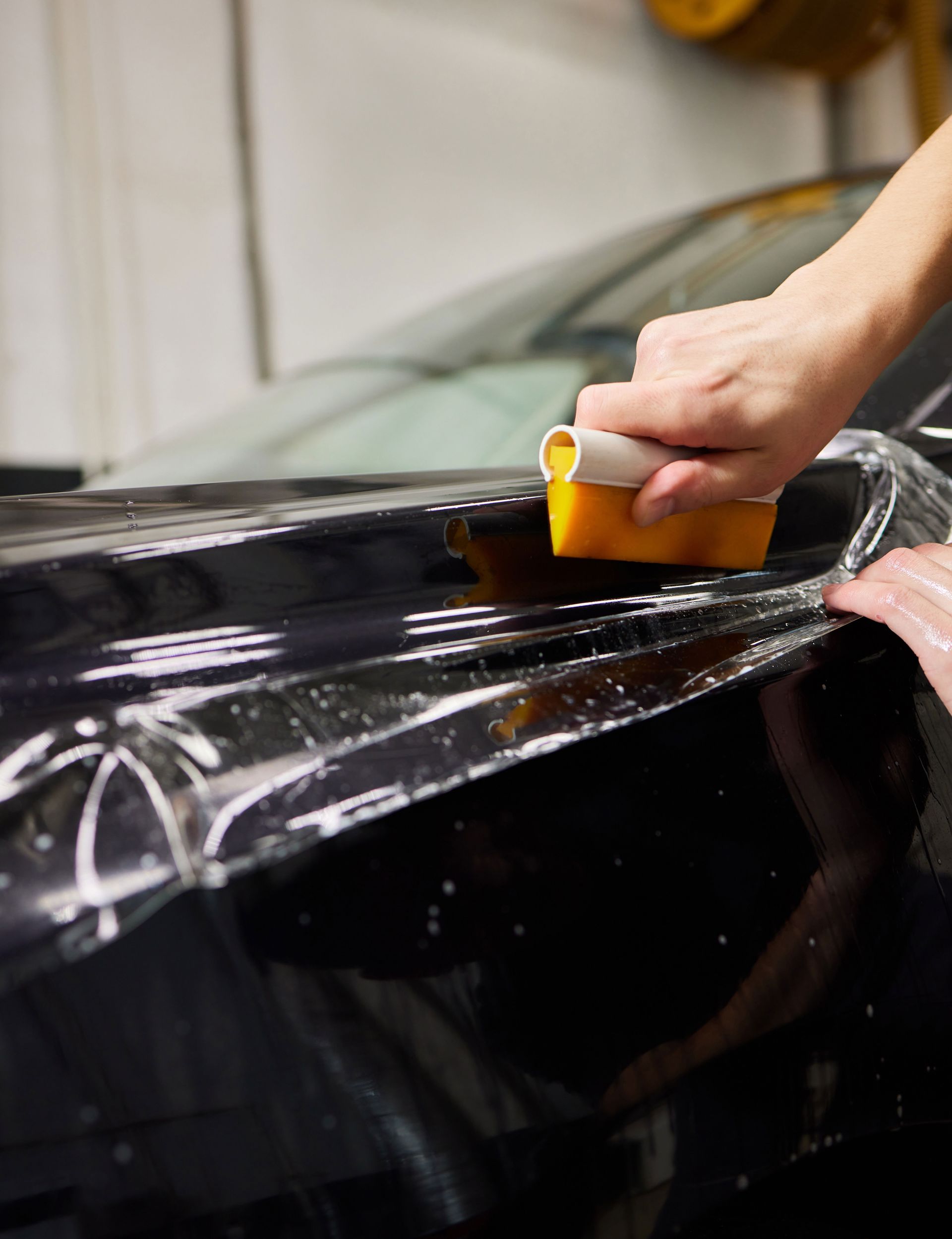 A person uses a yellow squeegee to smooth a clear protective film onto the black hood of a car.