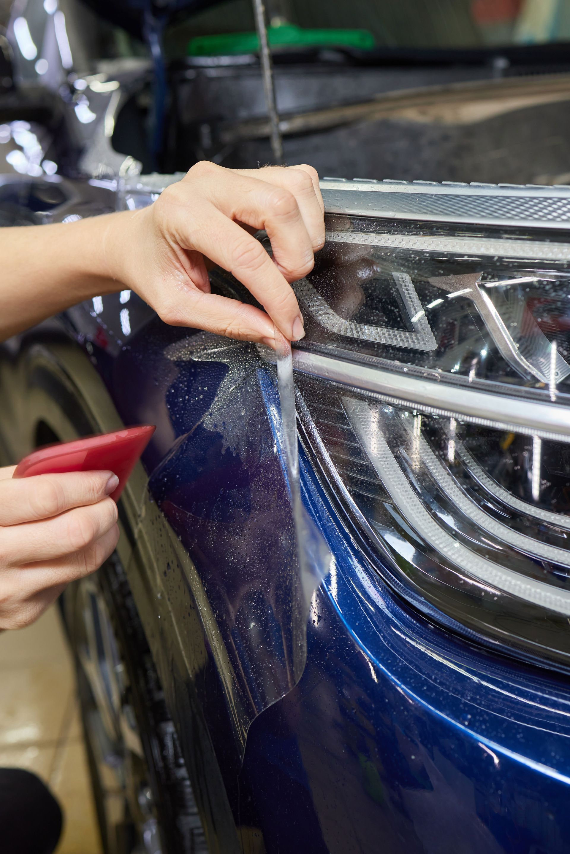 A person applies a clear paint protection film to the front corner of a dark blue car near the headlight using a squeegee.