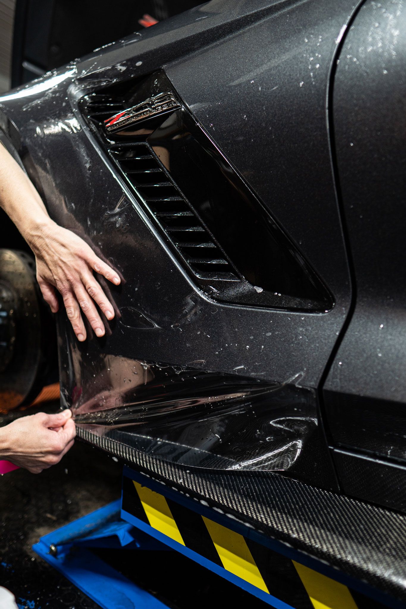 A person applies a clear protective film to the dark, vented fender of a vehicle.