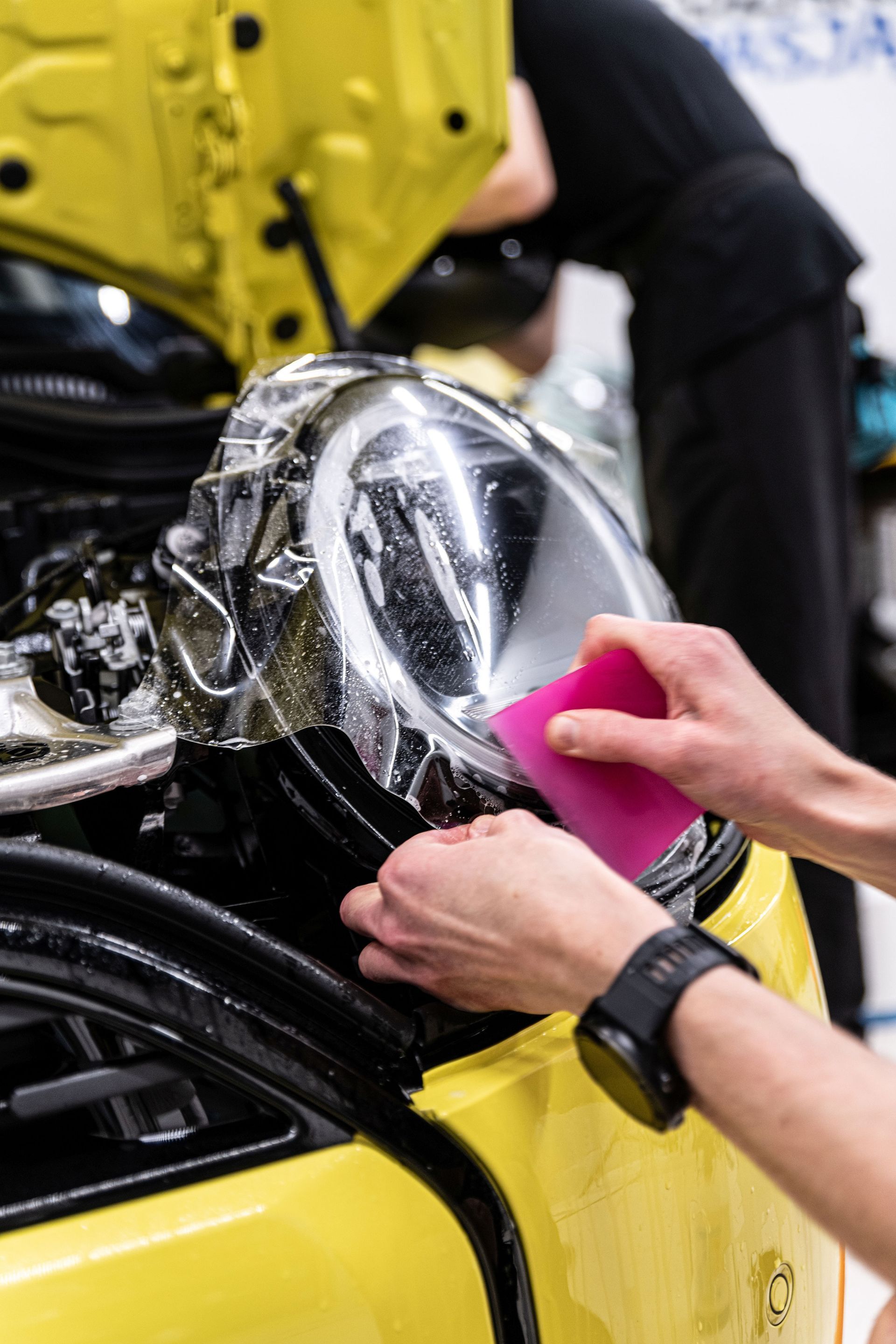 A technician applies a protective film to a yellow car's headlight using a pink squeegee in a workshop.