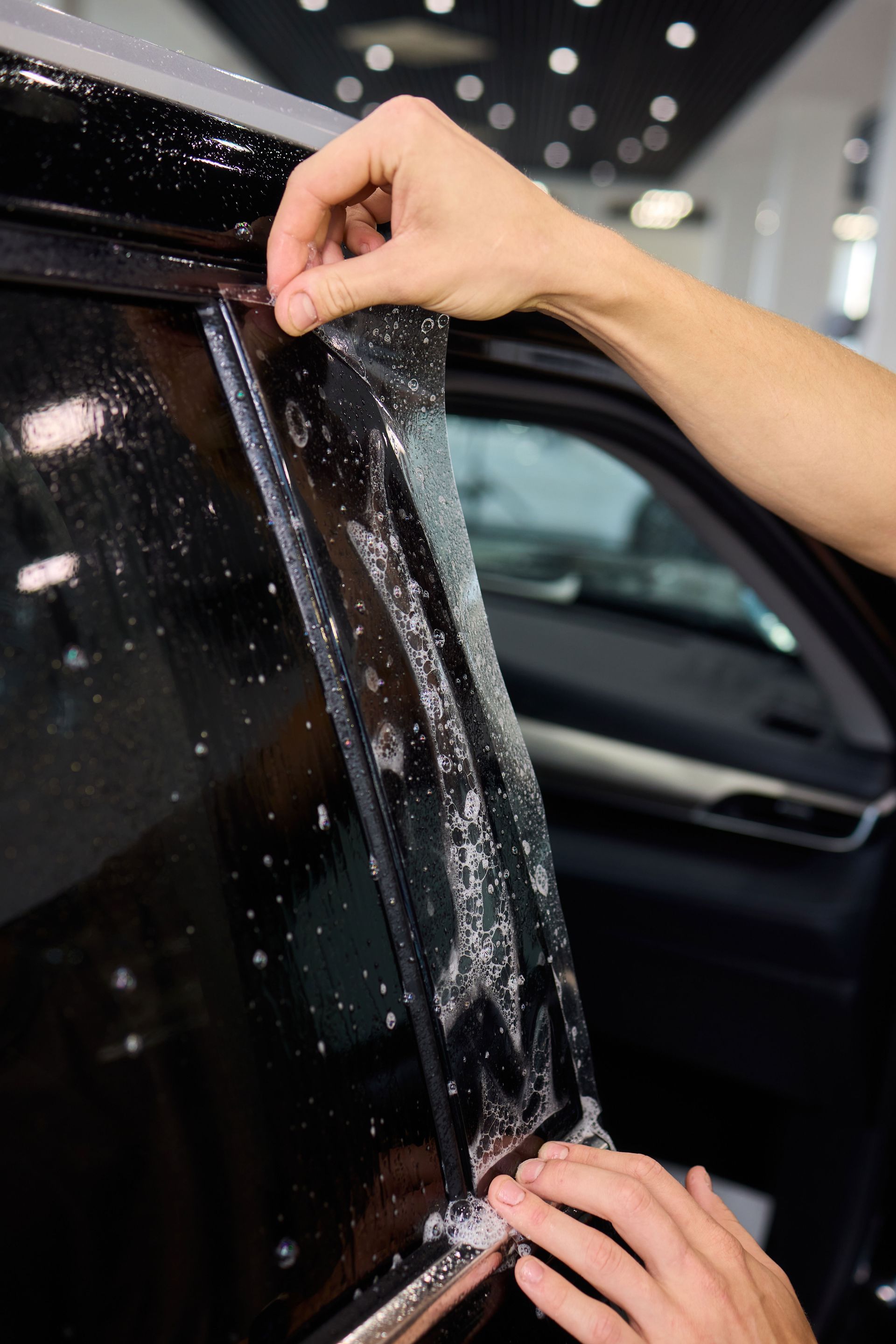 Hands applying a clear adhesive film to a car window using a soapy solution in a garage setting.