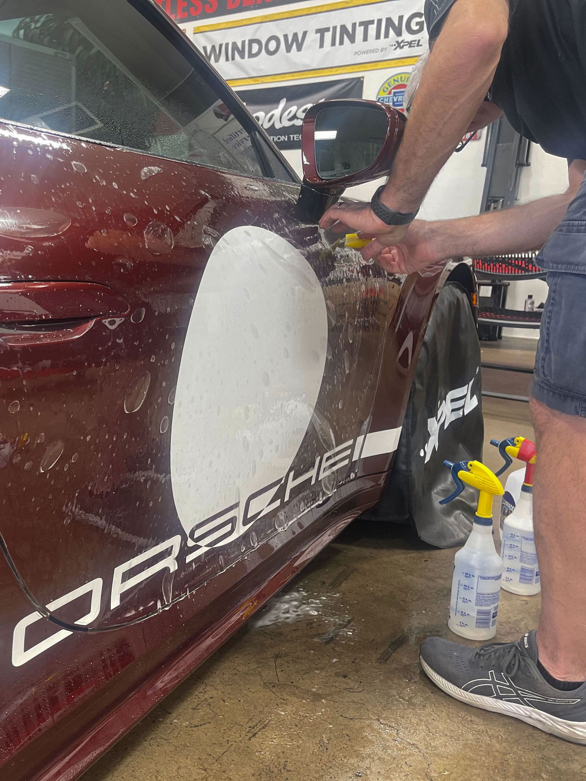 Man applying film to a red Porsche. The car has white racing stripes. Window tinting shop setting.