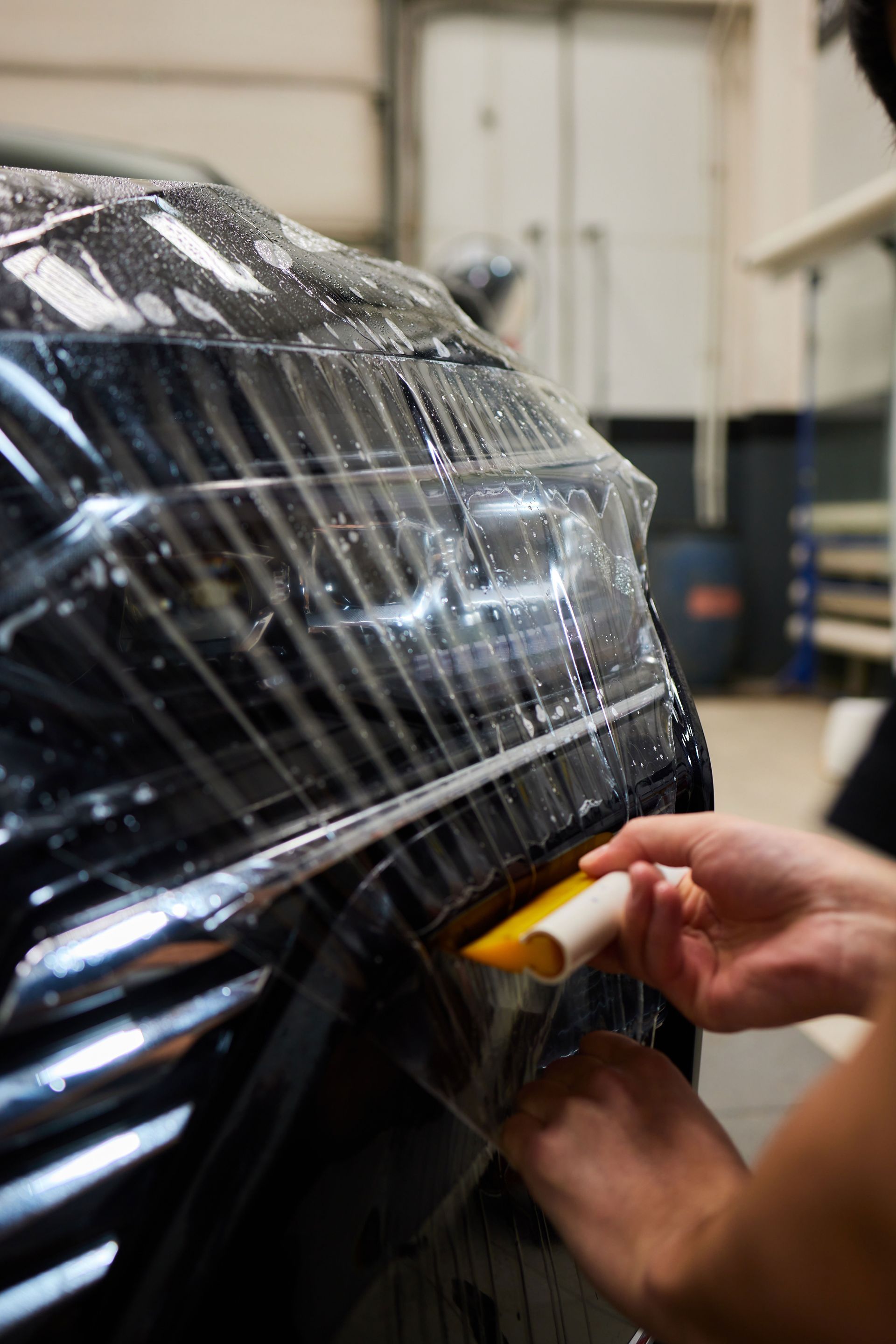 A close-up view of a person using a yellow squeegee to apply clear protective film to the front bumper of a black car.