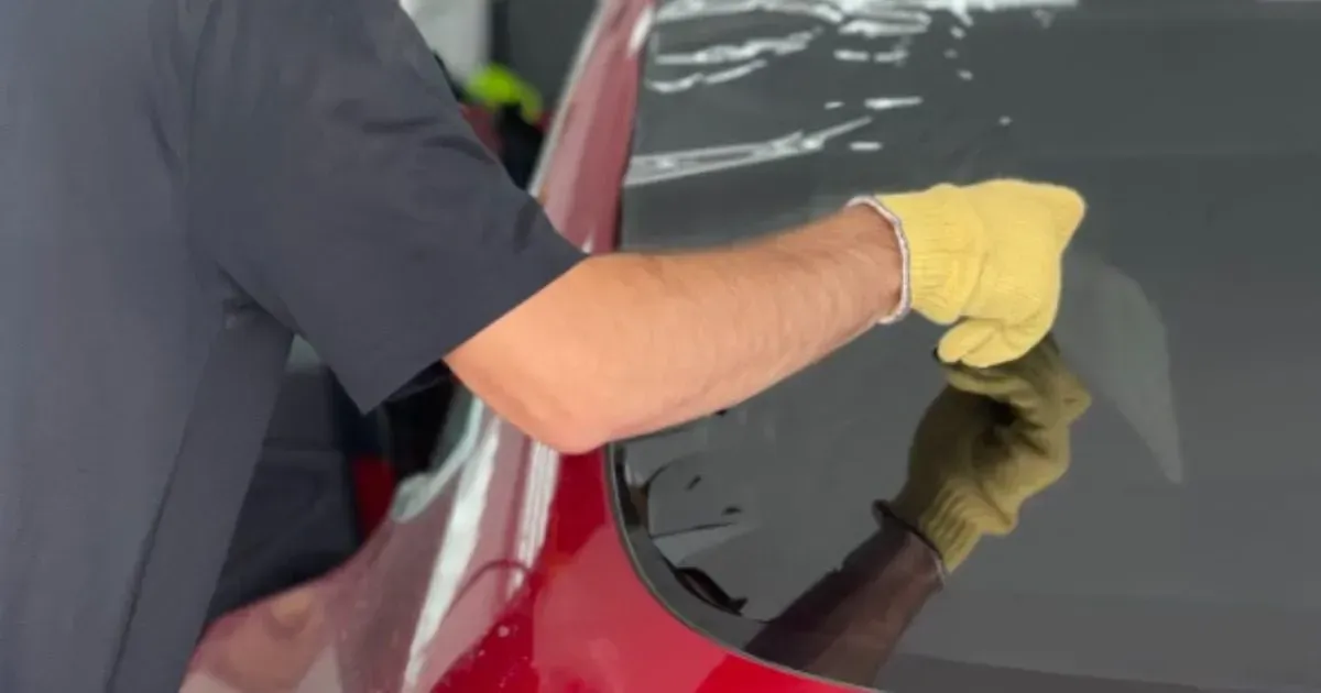 A person wearing a yellow work glove smooths a dark window tint film onto the rear glass of a red car.