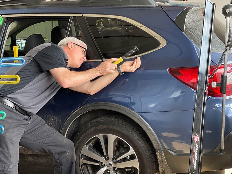 A person in a gray uniform uses a specialized tool to perform paintless dent repair on the side of a blue SUV.