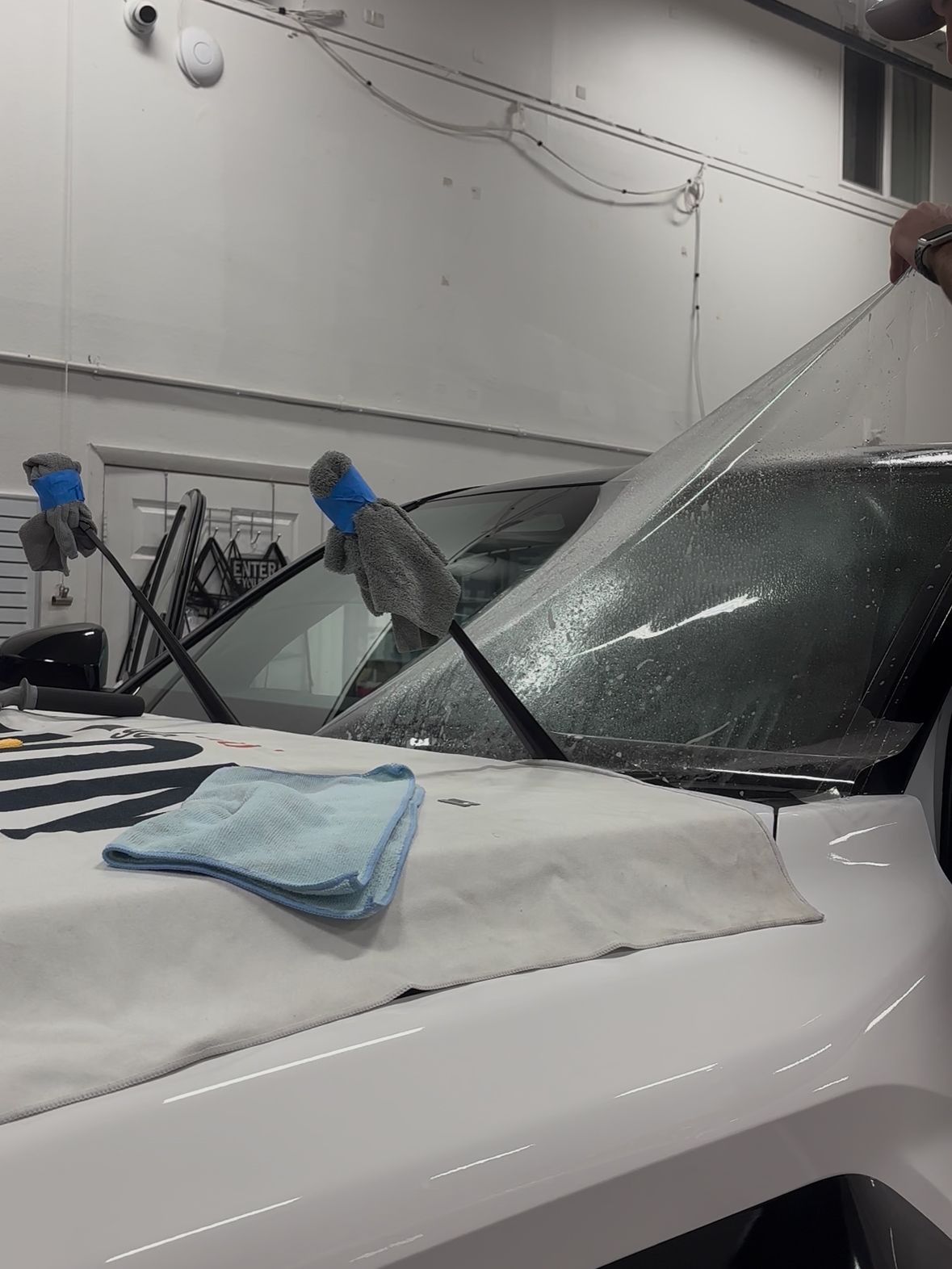 A person applying a clear protective film onto a vehicle windshield in an indoor shop setting.
