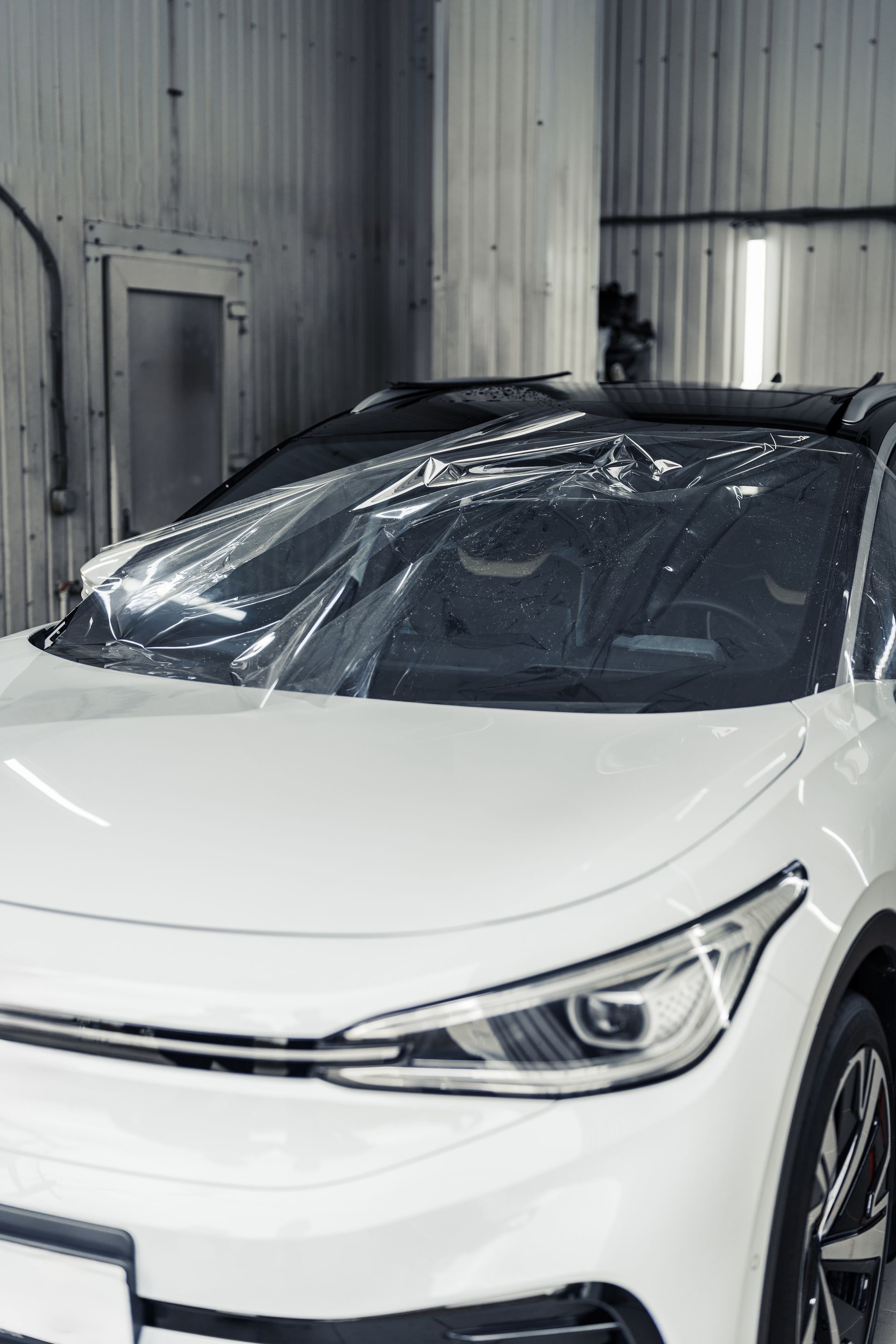 A white car with a protective plastic film covering its windshield sits inside an auto shop.