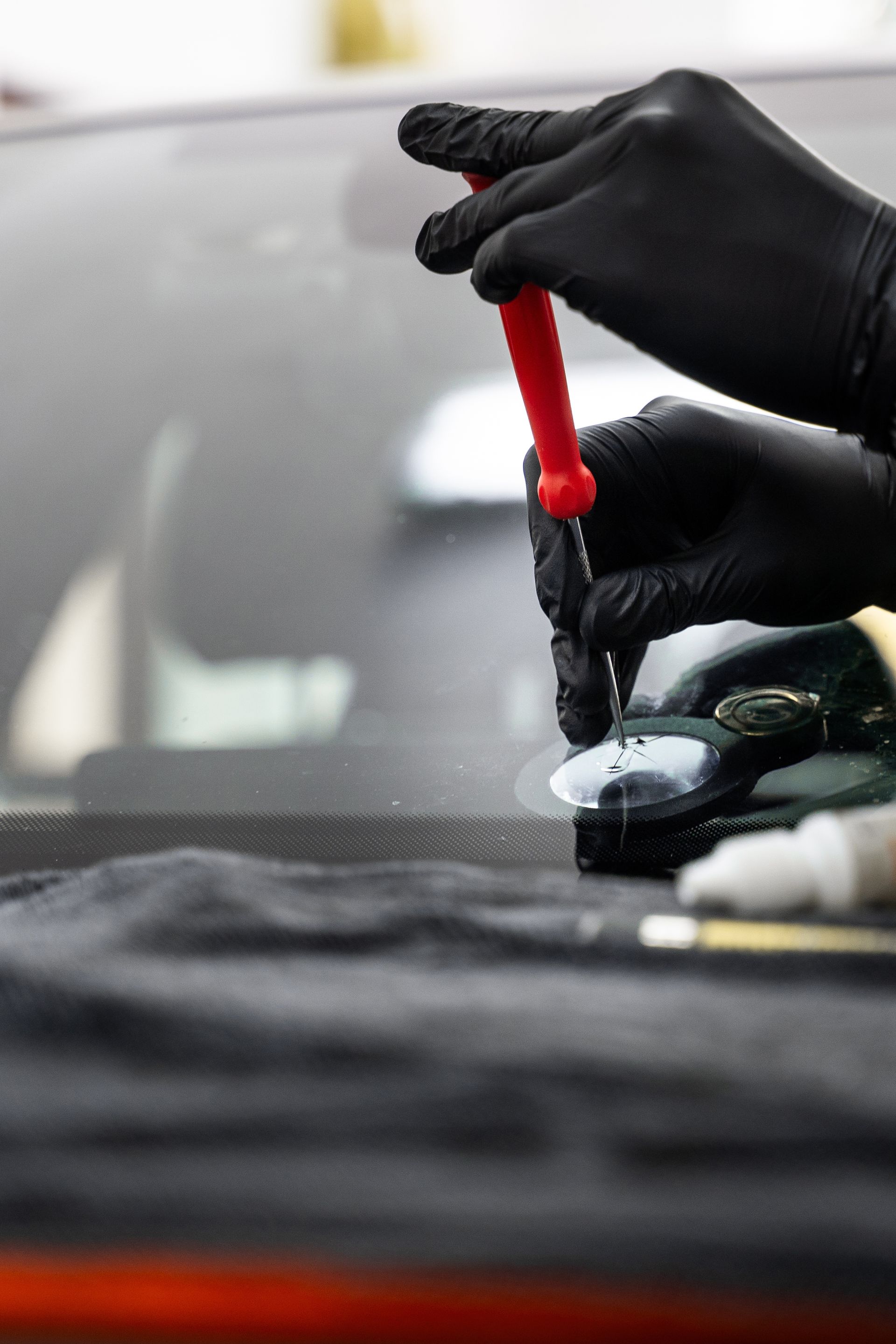 Gloved hands use a red-handled tool to repair a small chip in a car windshield.