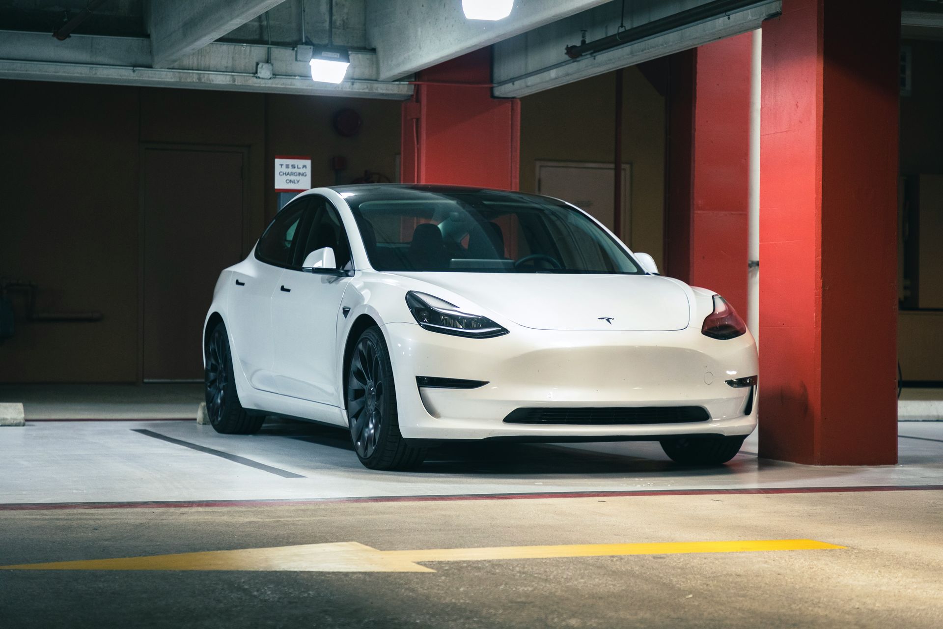 A white Tesla Model 3 parked in a brightly lit indoor parking garage with red pillars.