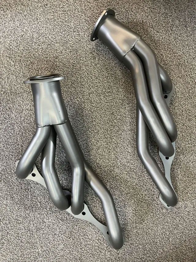 Two metallic, dark gray automotive exhaust headers for a V8 engine, laid out on a carpeted floor.