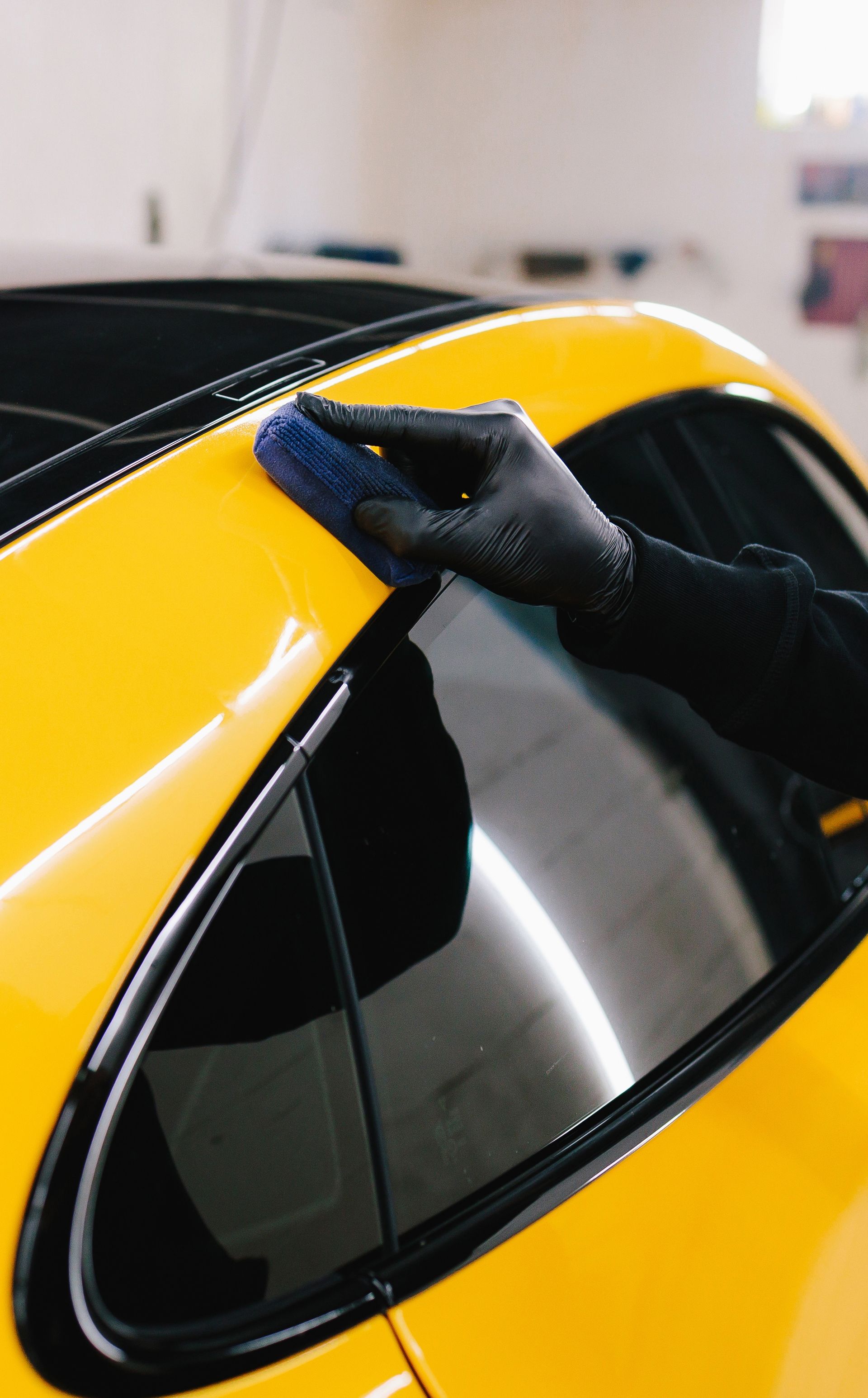 A person in black gloves uses a blue applicator pad to apply a coating to the yellow roof pillar of a car.
