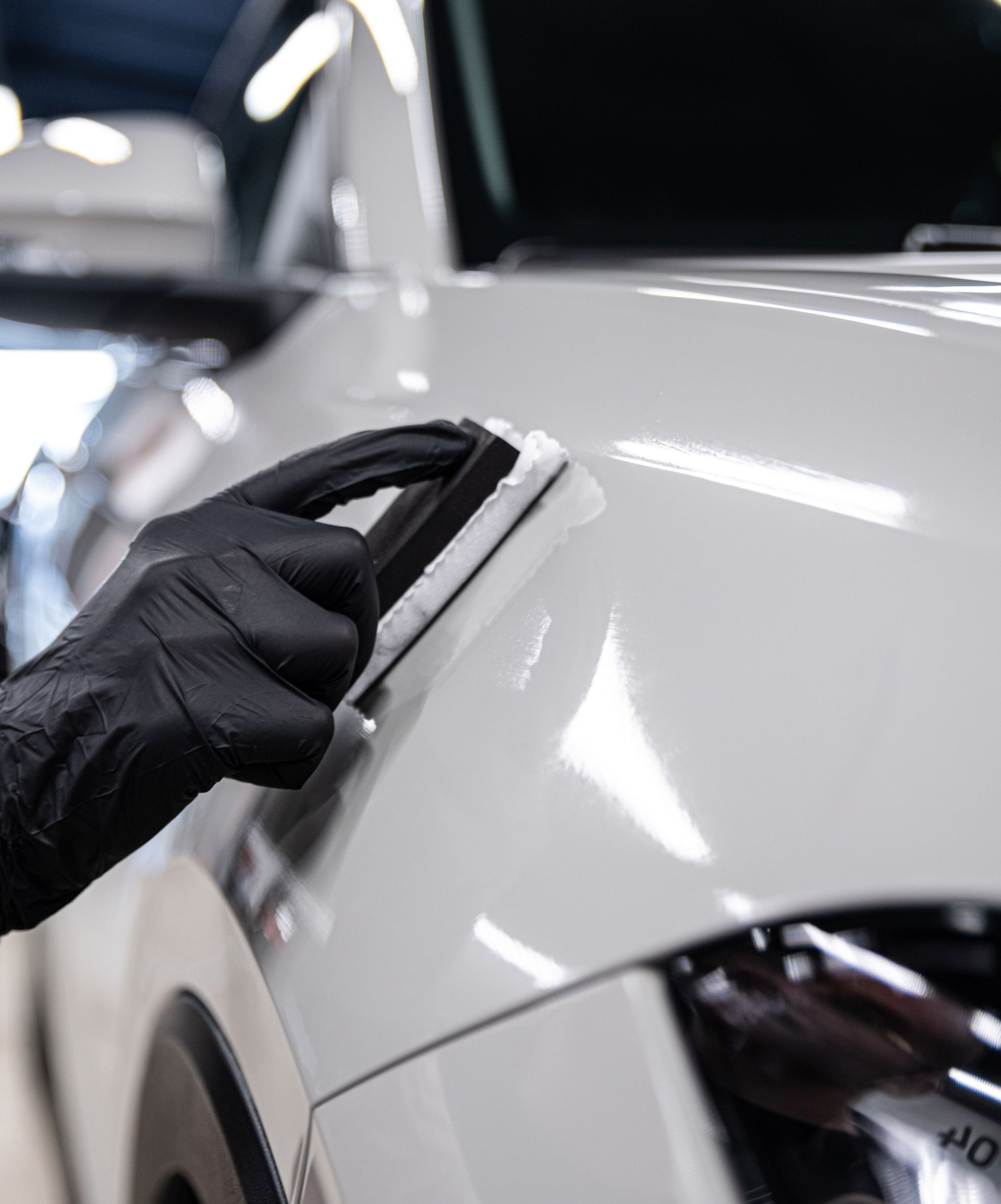 A person wearing black gloves uses an applicator pad to apply ceramic coating to a glossy white car body panel.