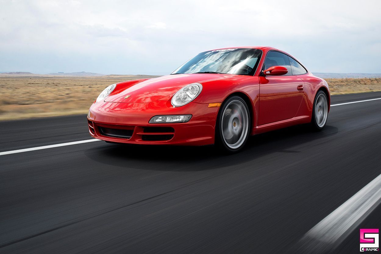 A red Porsche 911 coupe driving on an open, paved road against a clear, expansive landscape.
