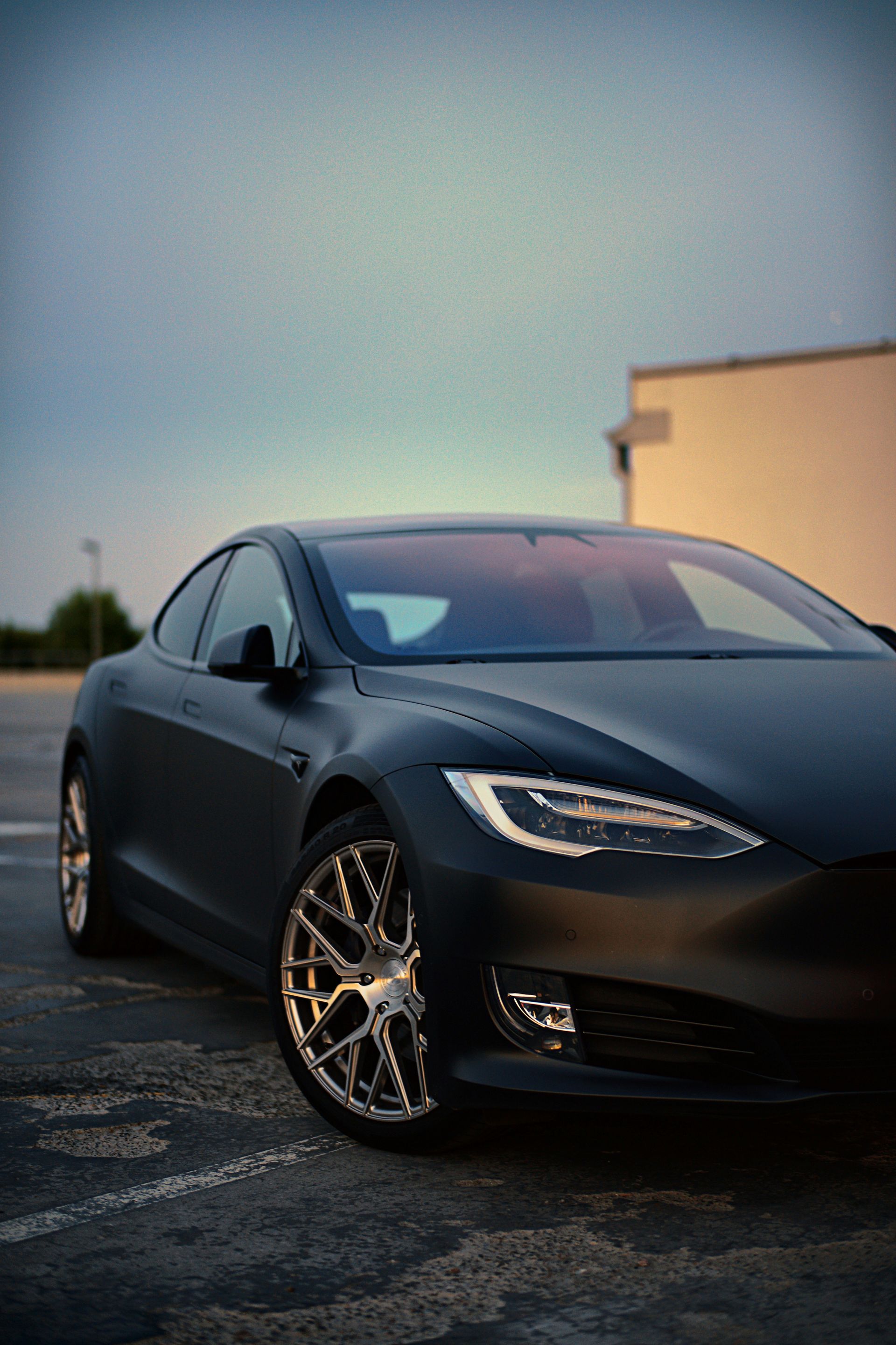 A matte black Tesla Model S with custom silver wheels parked on an asphalt lot at dusk.
