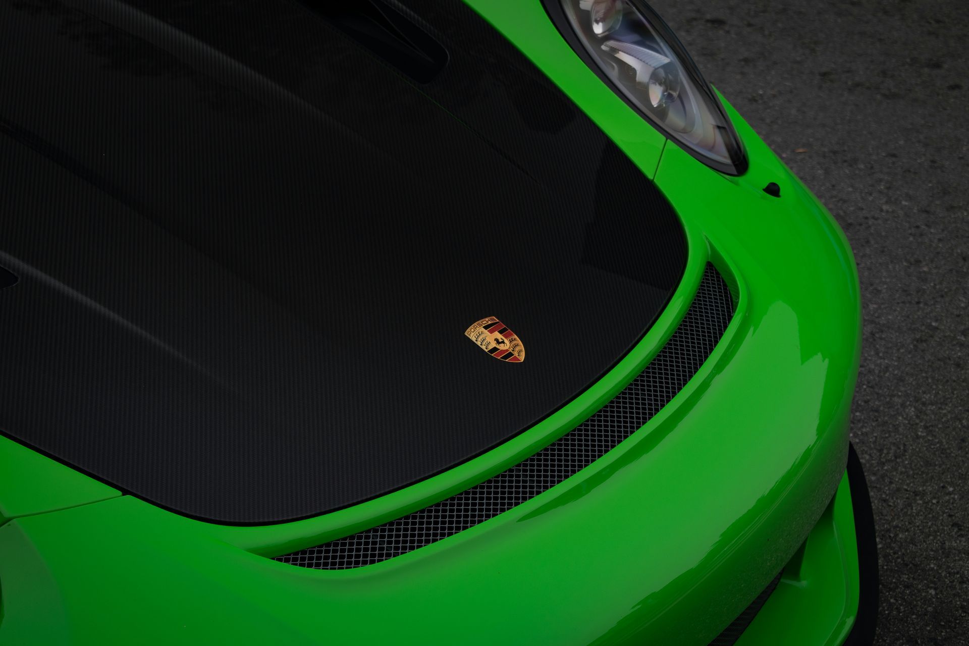 A bright green Porsche with a carbon fiber hood and the iconic golden crest emblem on the front.