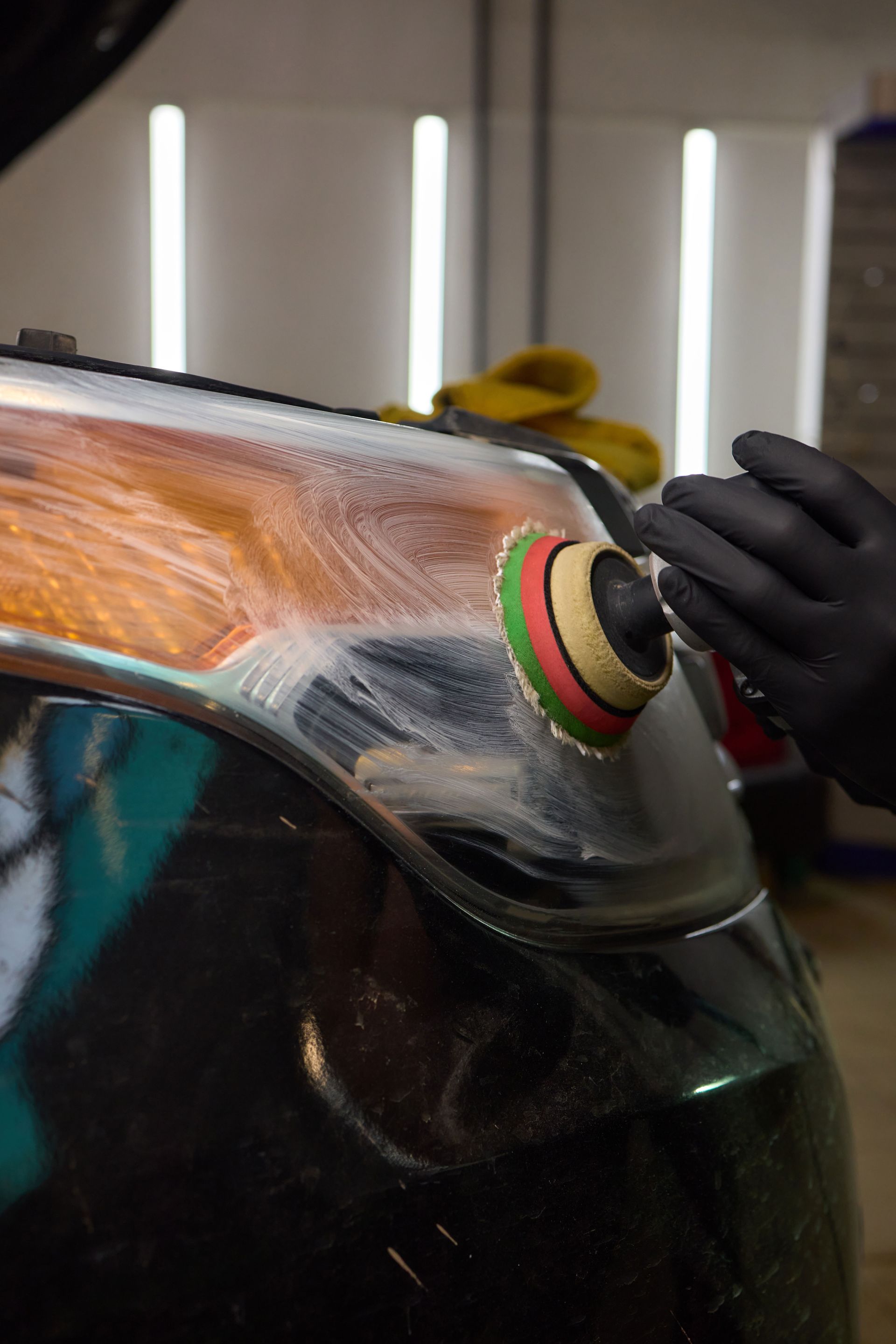 A gloved hand uses a rotary polisher with a colorful foam pad to buff and clear a car headlight.