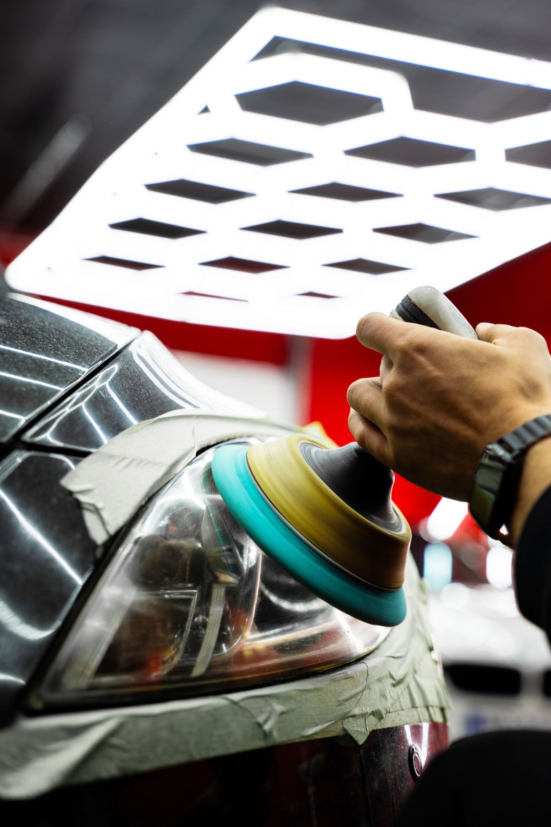 A person uses a rotary buffer with a blue foam pad to polish a car headlight, reflected by a grid light above.