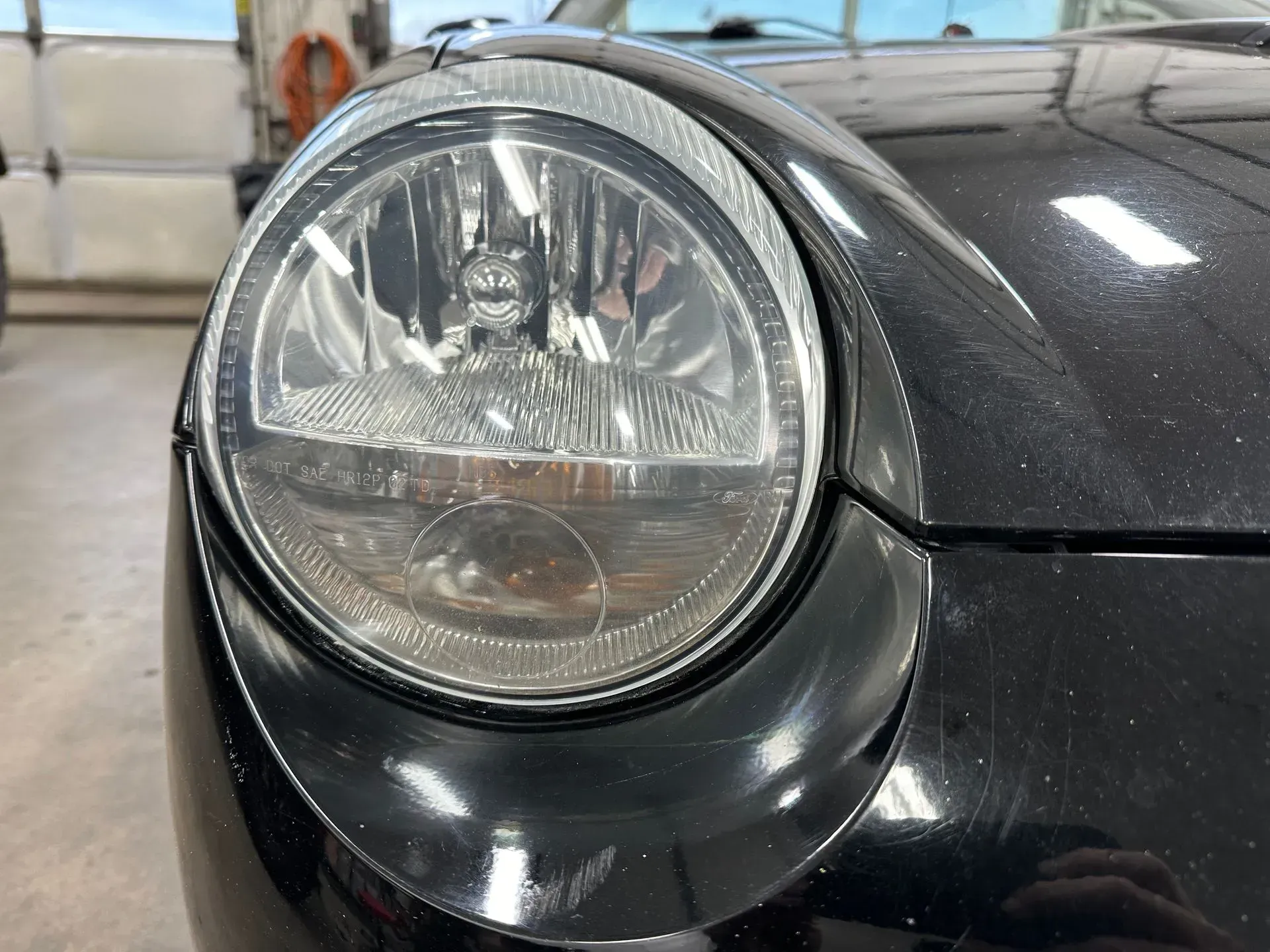 A close-up of a circular car headlight on a black vehicle, positioned in a garage setting with reflections on the surface.