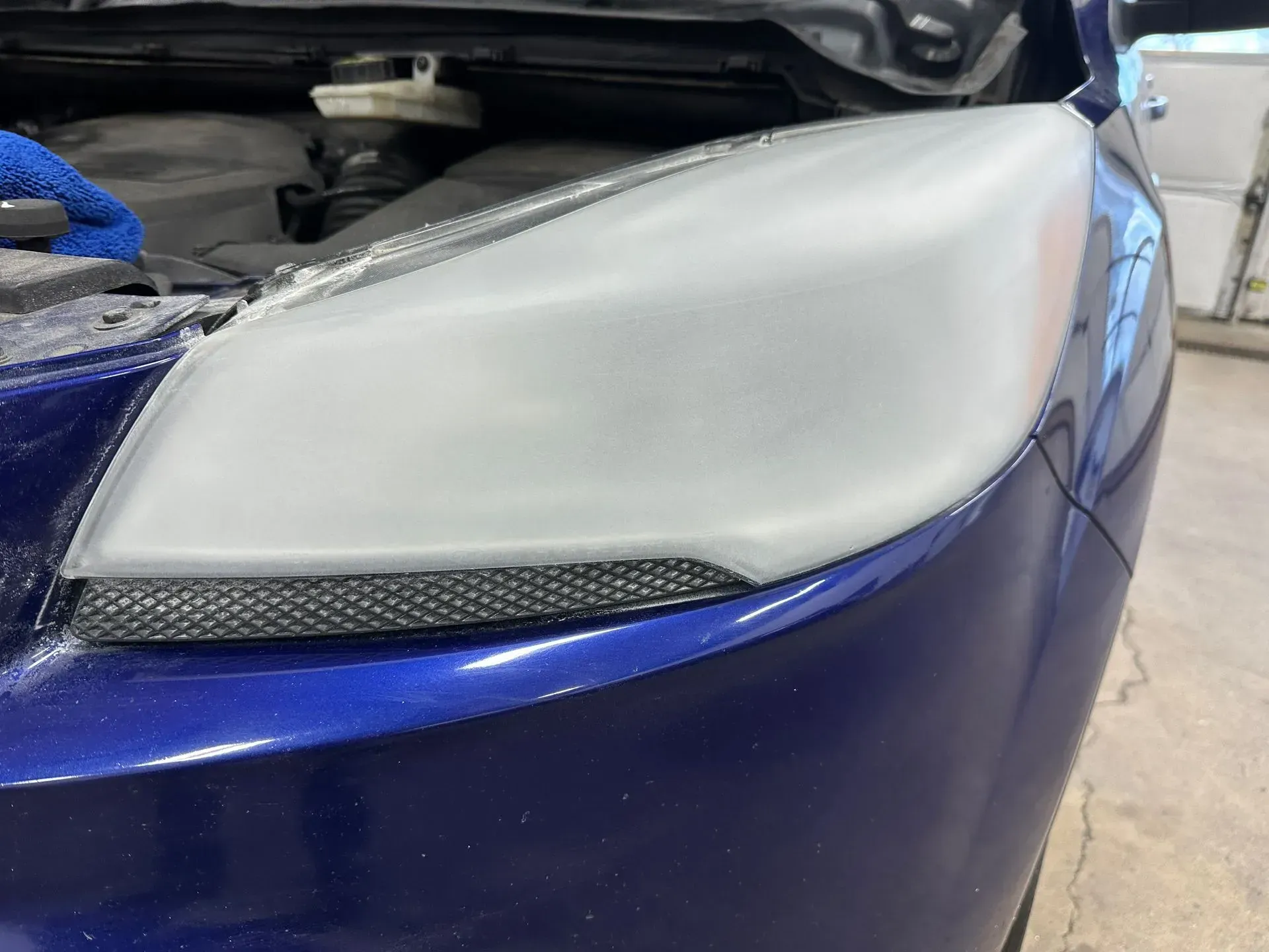 A blue car's headlight assembly that has been sanded down, appearing matte and opaque during a restoration process.