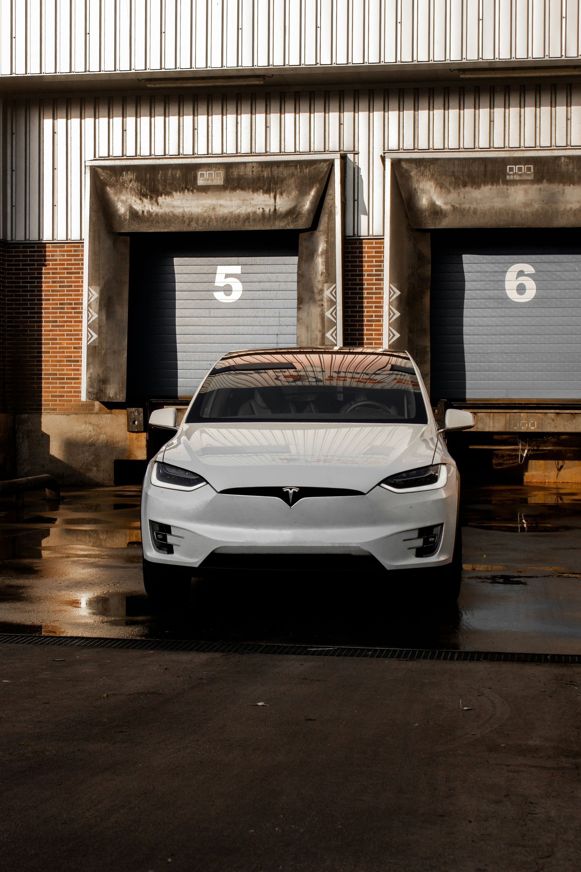 A white Tesla Model X parked in front of two industrial shipping bays labeled 5 and 6.
