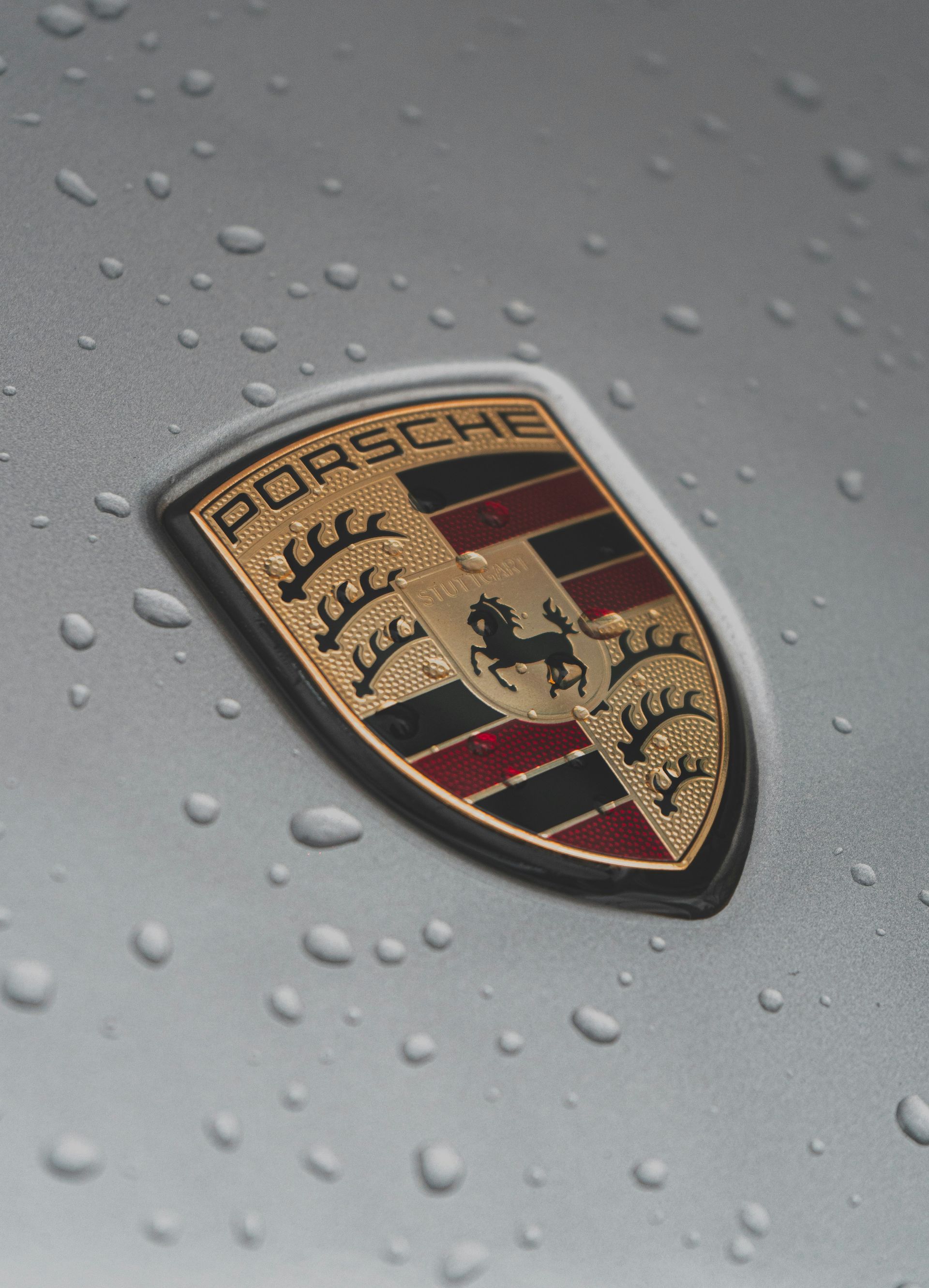 The Porsche logo emblem on a silver surface covered in water droplets.