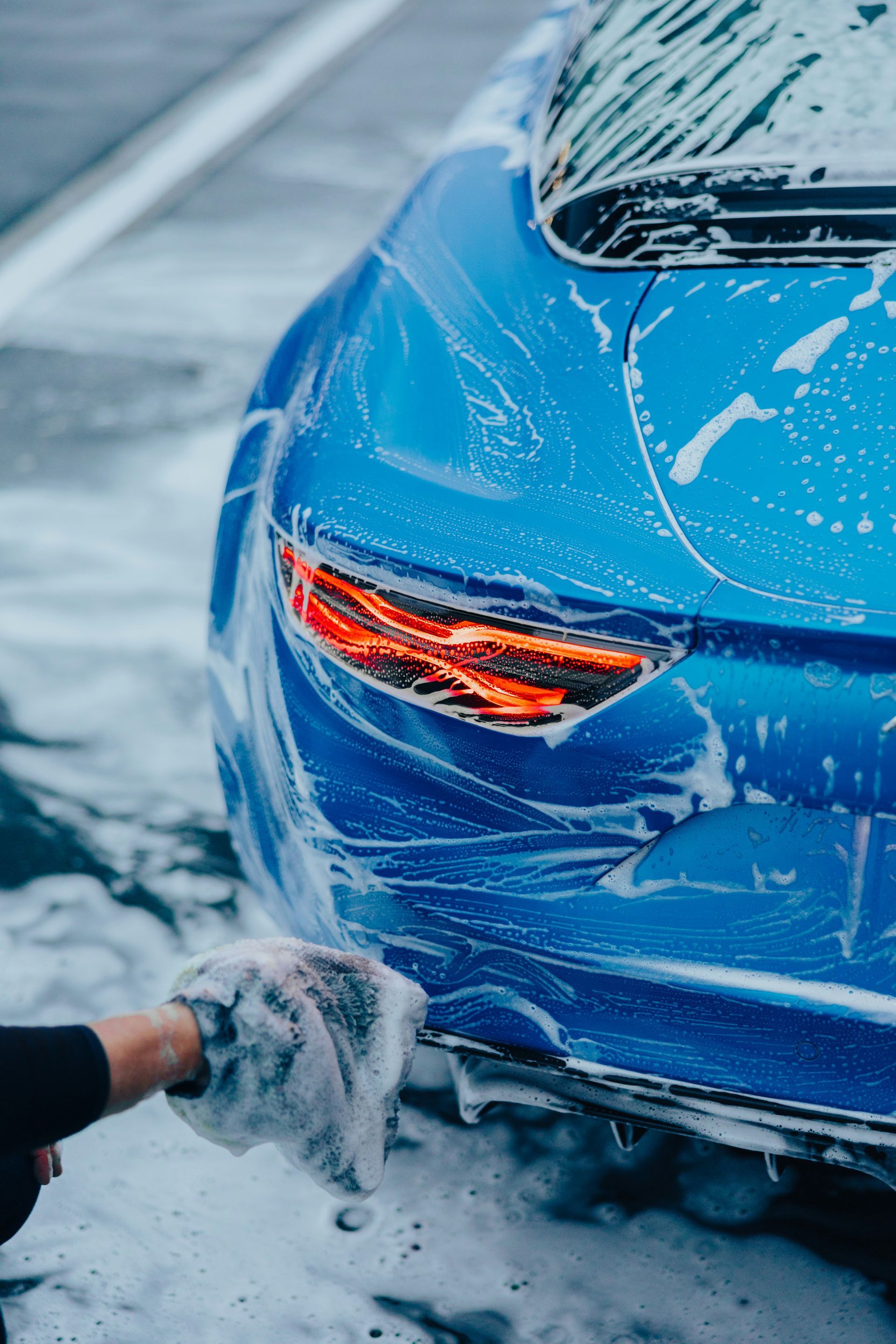 A person uses a grey wash mitt to soap up the rear section of a bright blue sports car.