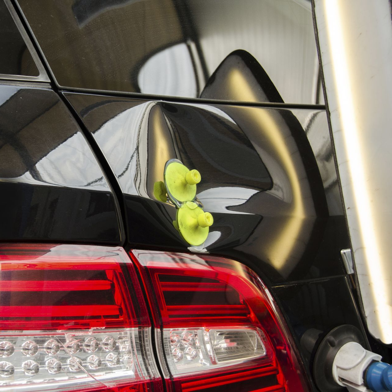 A technician uses a light board to repair a dent on the rear hatch of a dark-colored SUV in a bright auto repair shop.