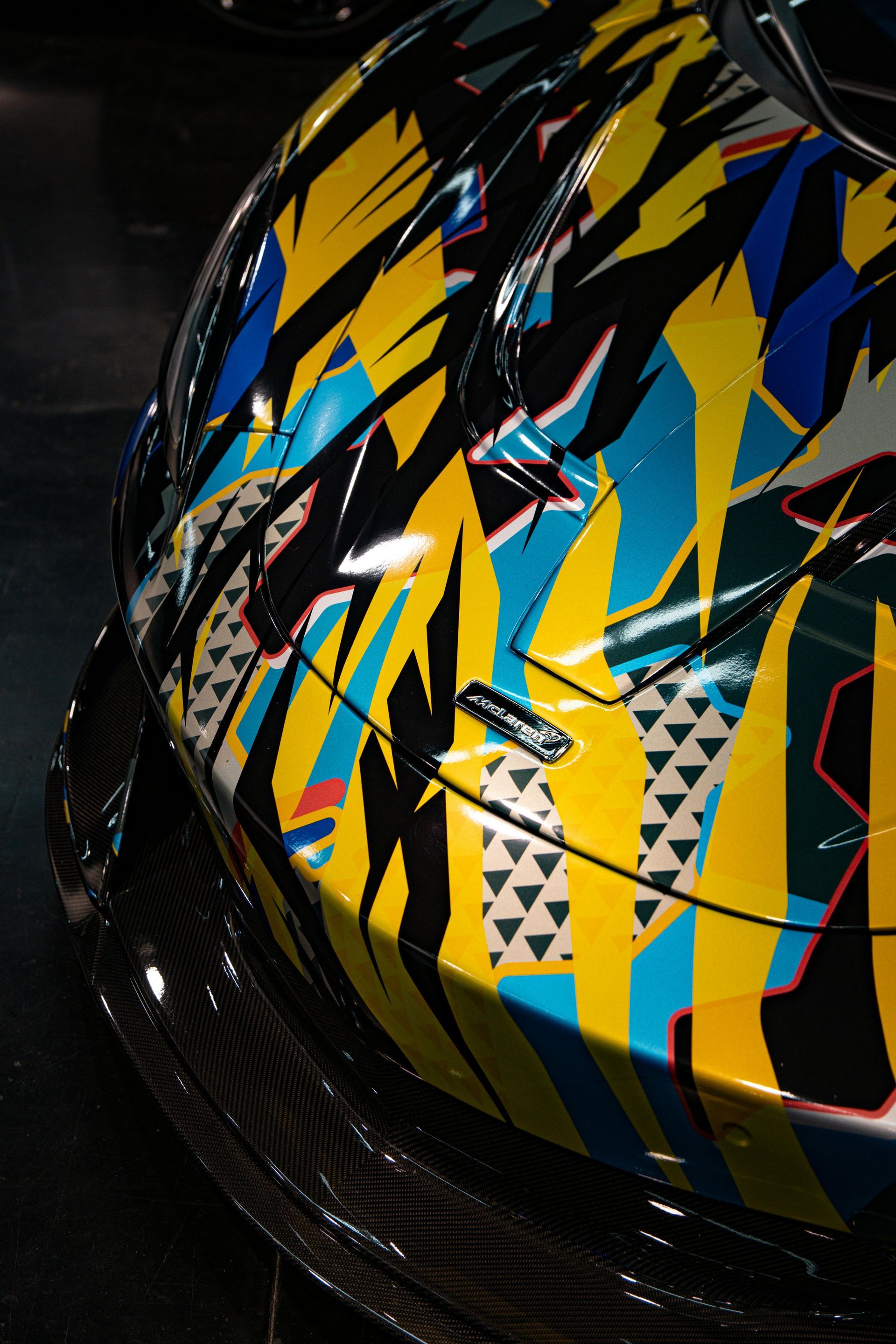 Close-up of a sports car hood featuring a vibrant yellow, blue, and black geometric graphic wrap and a carbon fiber bumper.