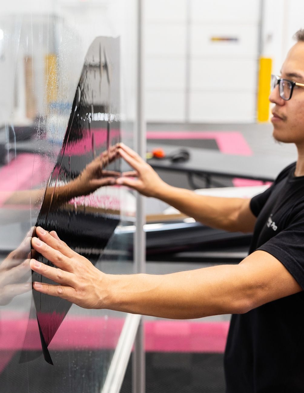 A person in a black t-shirt carefully applies a sheet of dark window tint to a clear pane.