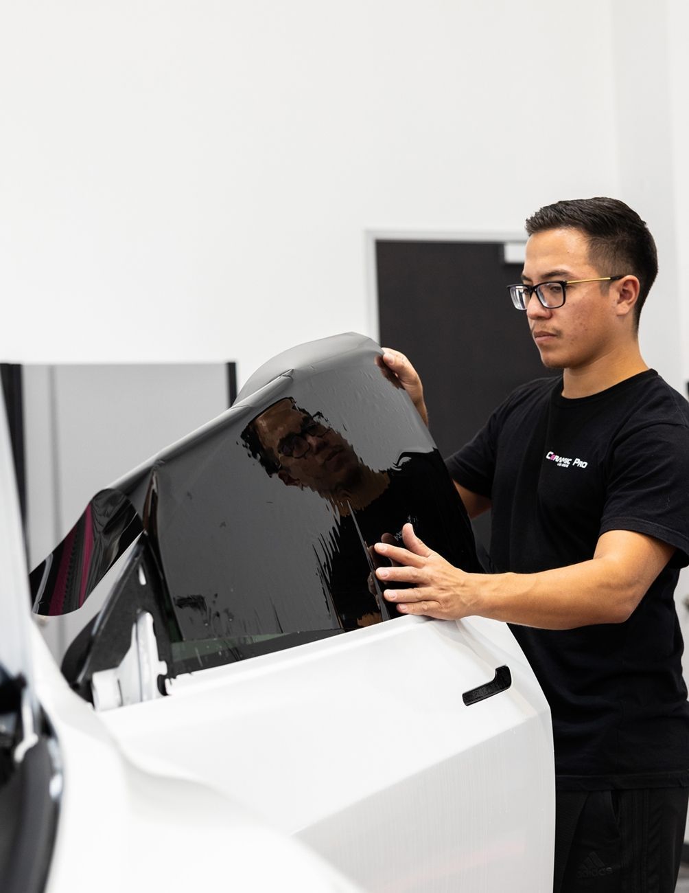 A person in a black t-shirt applying a dark window tint film to the car door of a white vehicle in a shop.