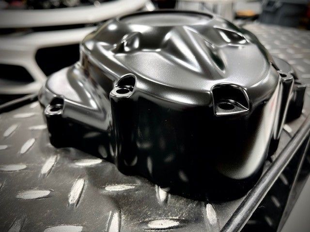 A matte black motorcycle engine cover rests on a diamond-plate metal surface in a workshop.