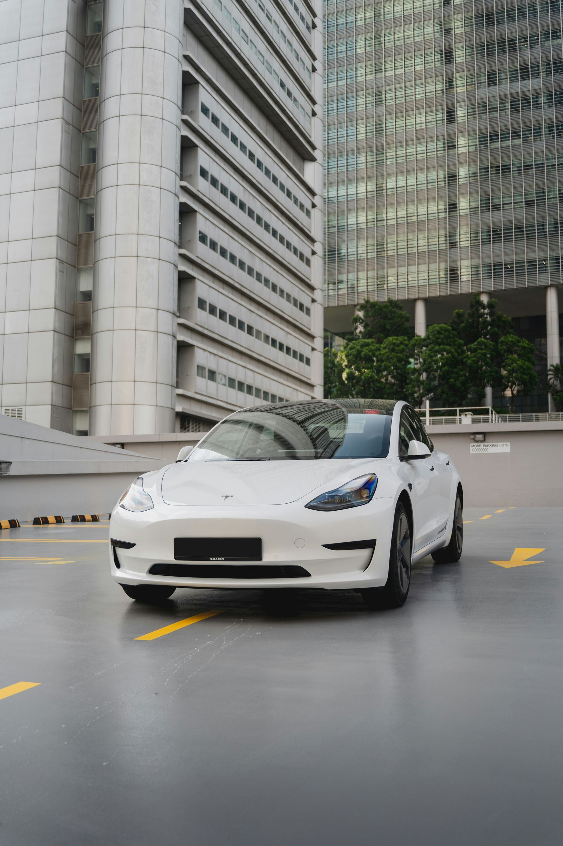 A white Tesla Model 3 parked on a concrete lot in front of modern high-rise buildings.