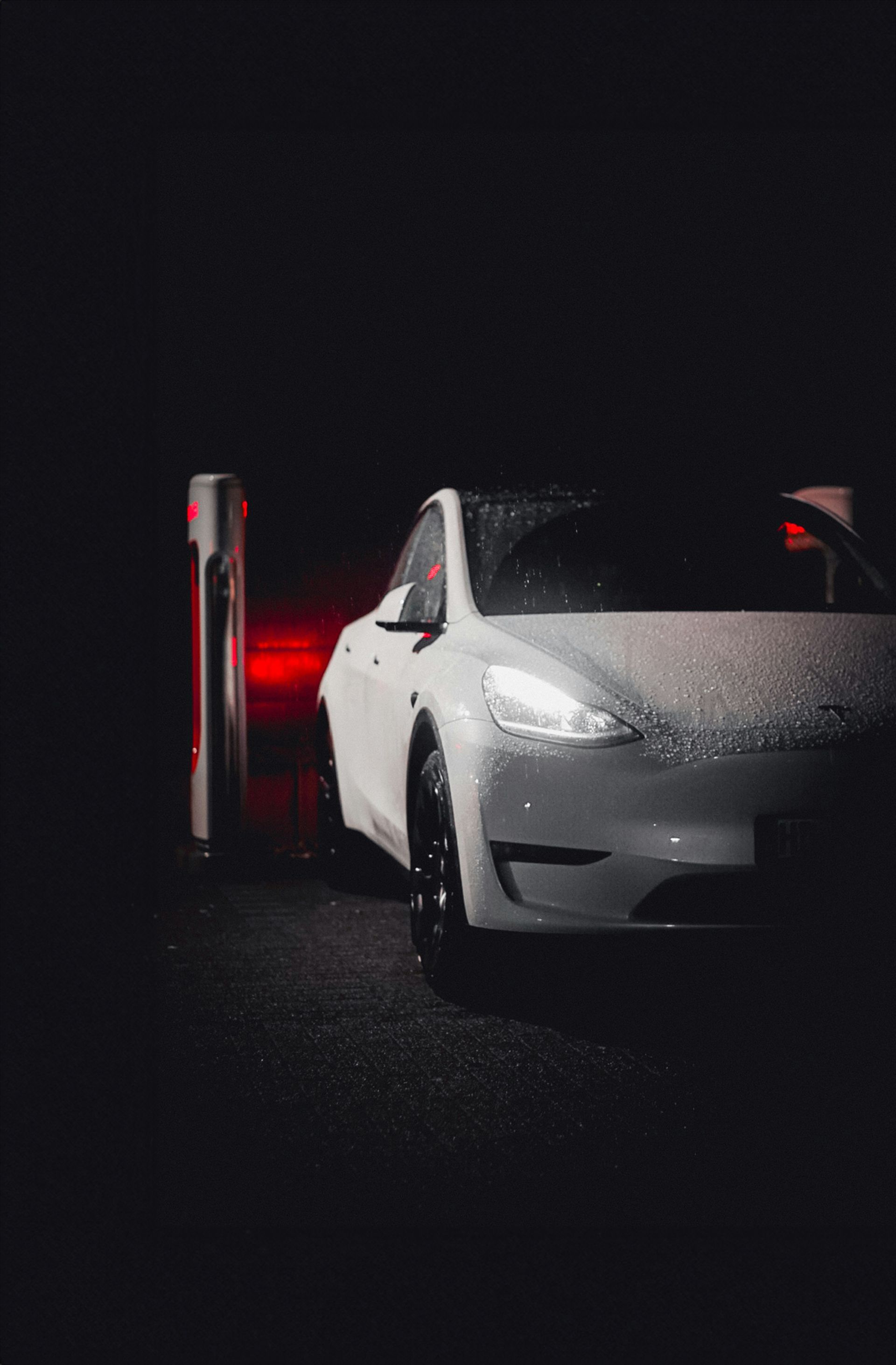 A white Tesla charging at a Supercharger station at night, covered in small raindrops.