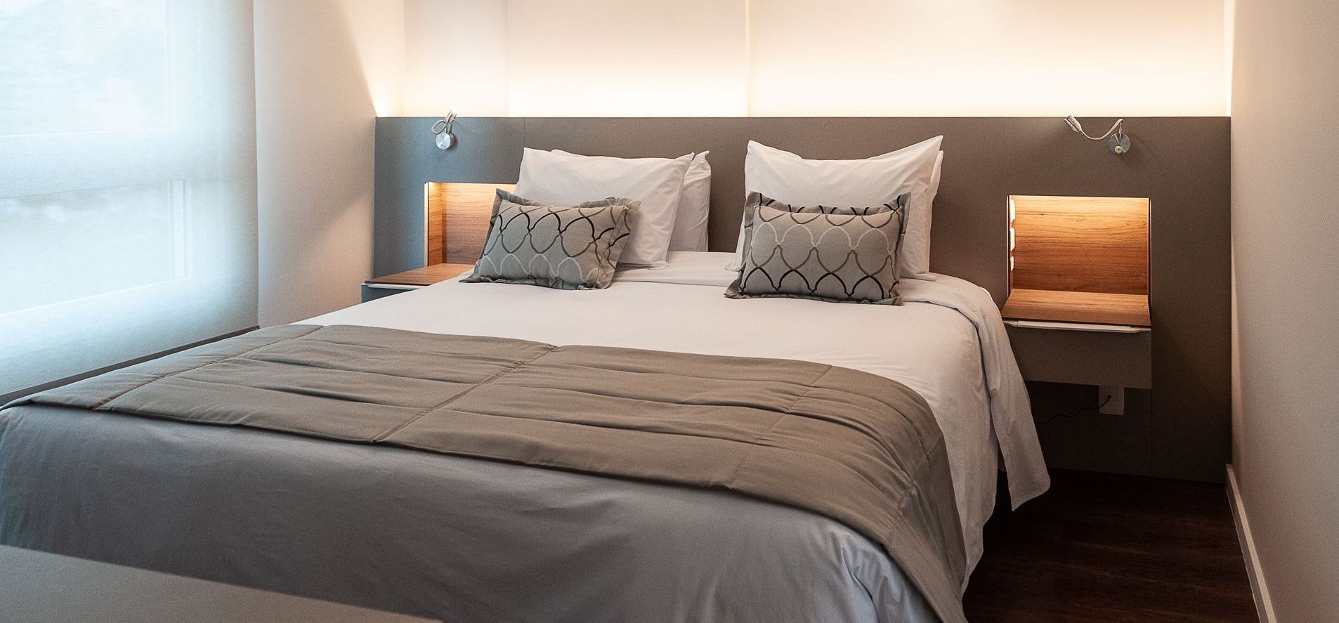 Quarto moderno com cama arrumada, roupa de cama cinza e iluminação aconchegante ao lado da cabeceira.
