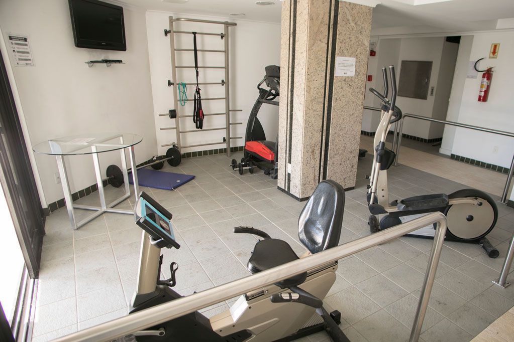 Sala de ginástica com bicicletas ergométricas, esteira e outros equipamentos de ginástica em piso acarpetado.