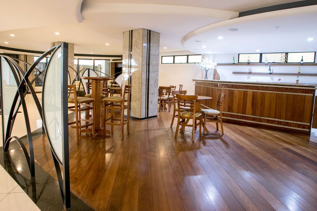 Interior de bar moderno e vazio, com piso de madeira, banquetas, mesas e balcão curvo branco.