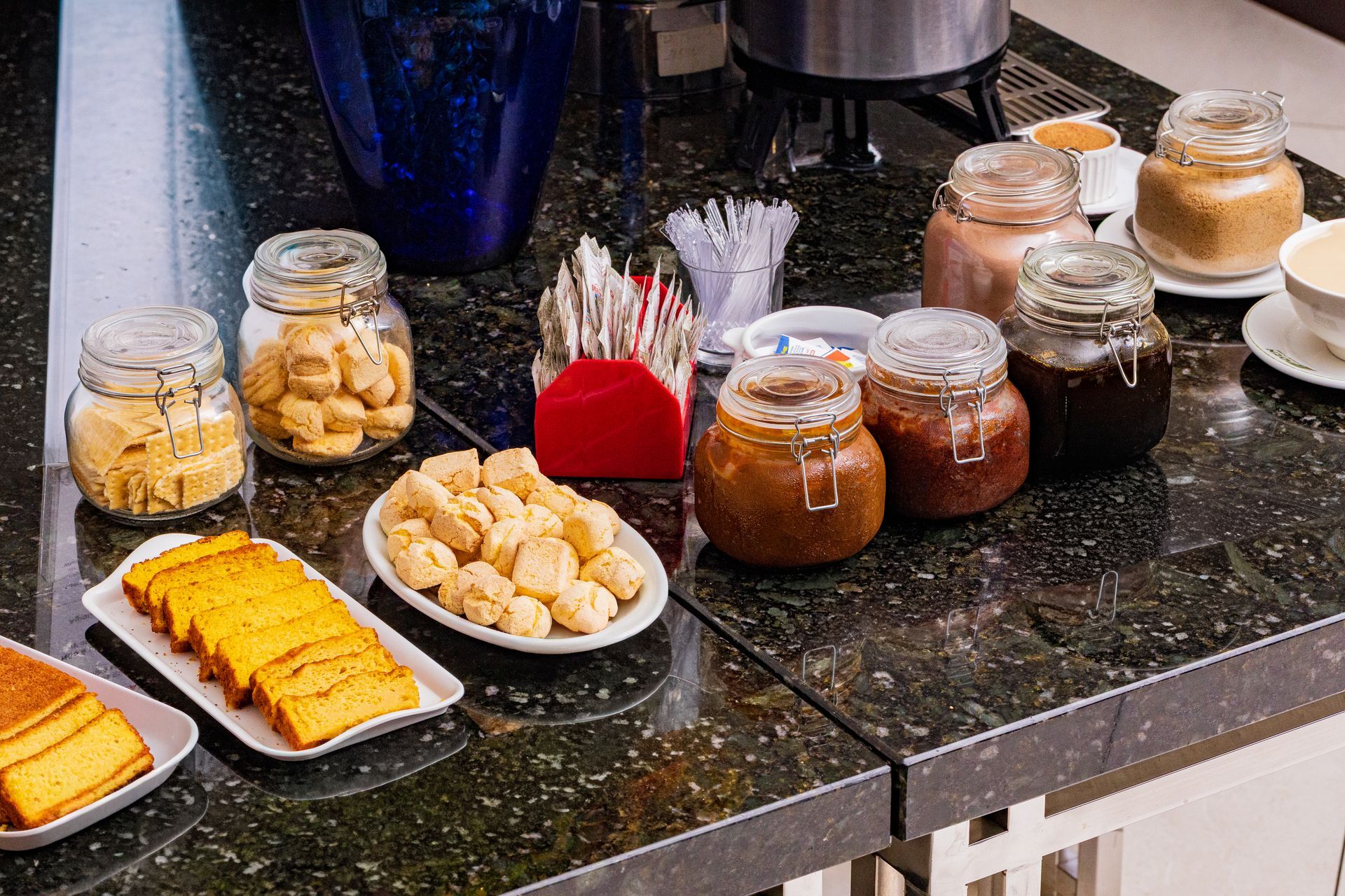 Buffet de café da manhã com potes de recheios, doces e fatias de bolo em uma bancada preta.
