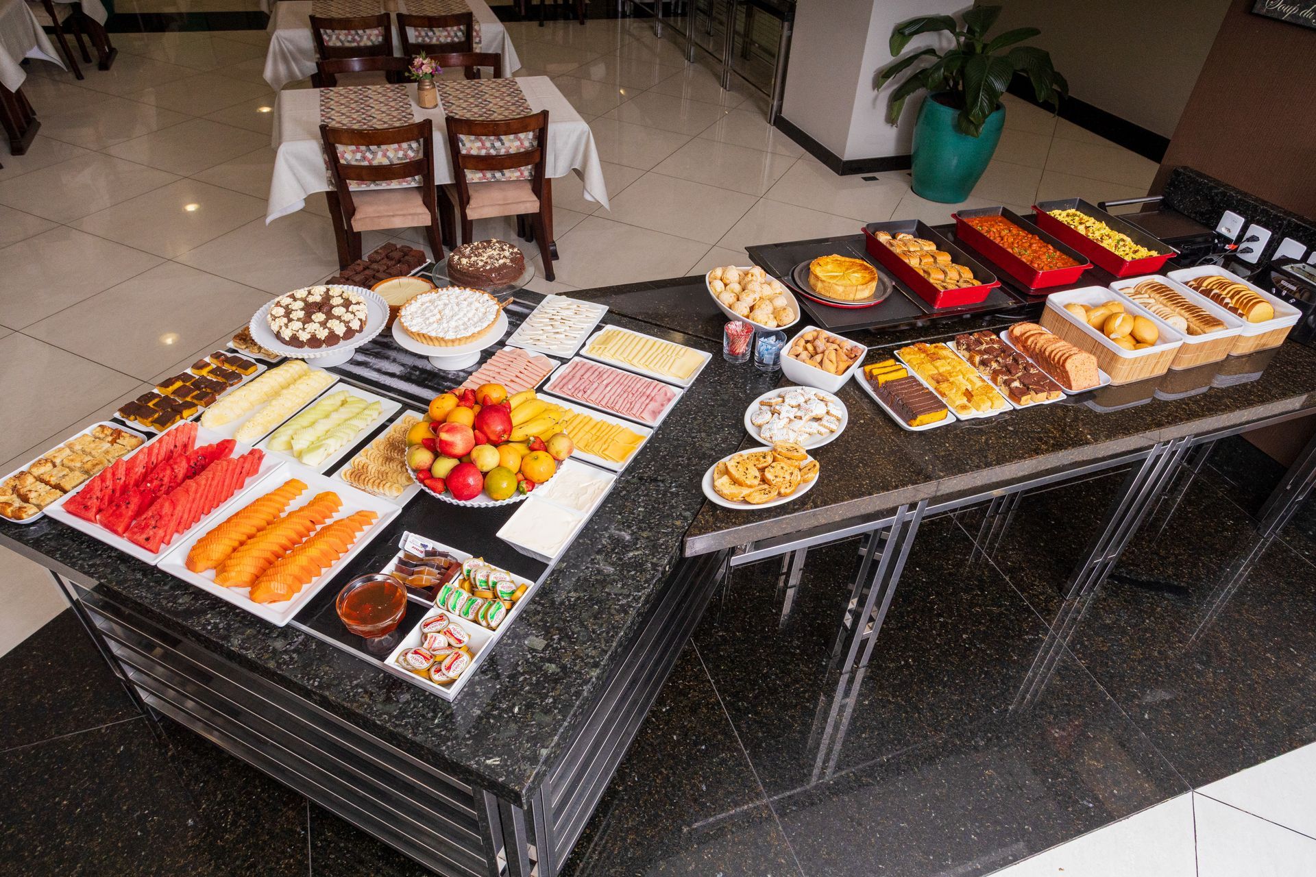 Mesa de bufê com sobremesas variadas, doces e frutas sobre toalhas de mesa pretas em uma sala de jantar.