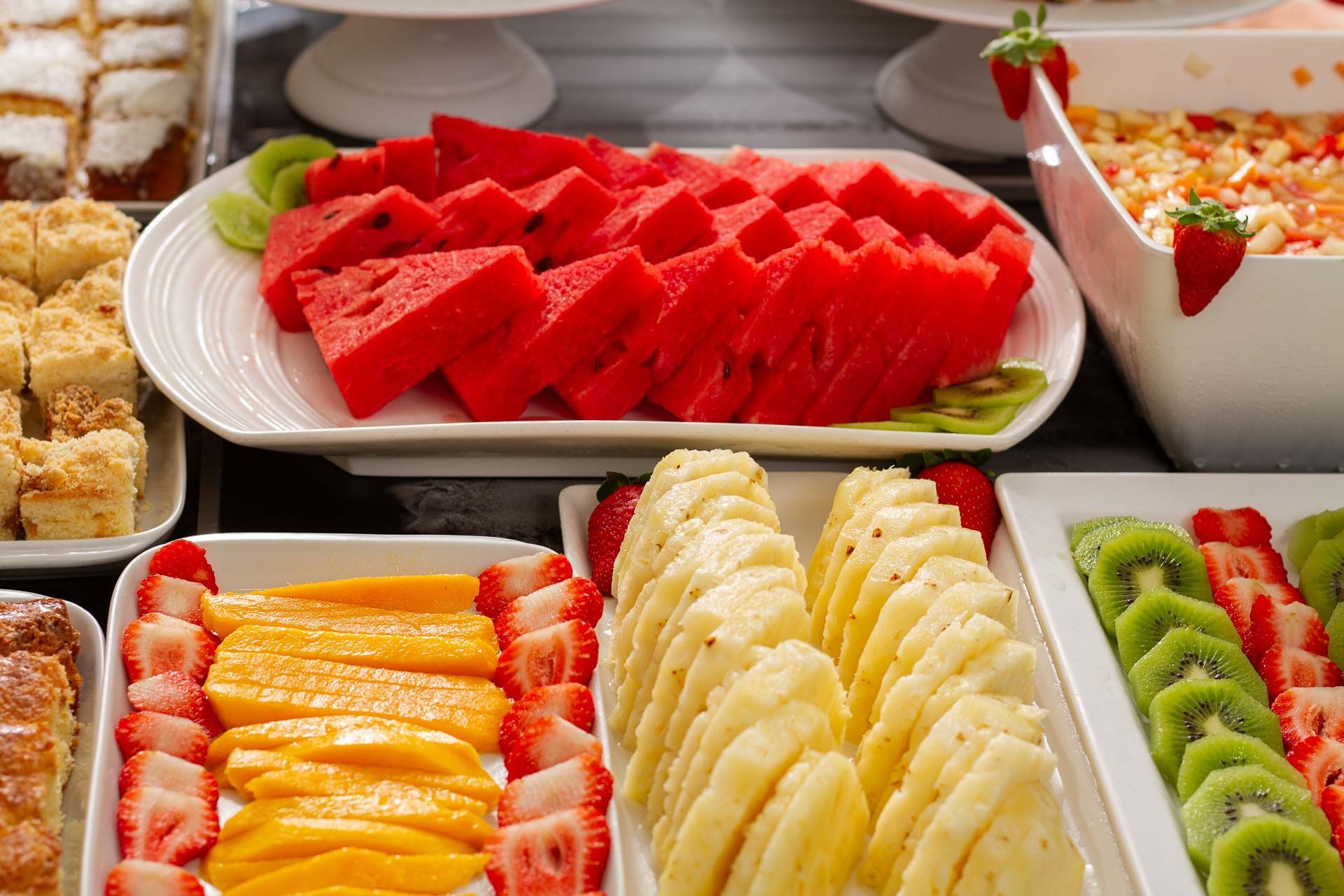 Frutas variadas fatiadas, incluindo melancia, abacaxi, manga, kiwi e morangos, dispostas em uma bandeja de bufê.