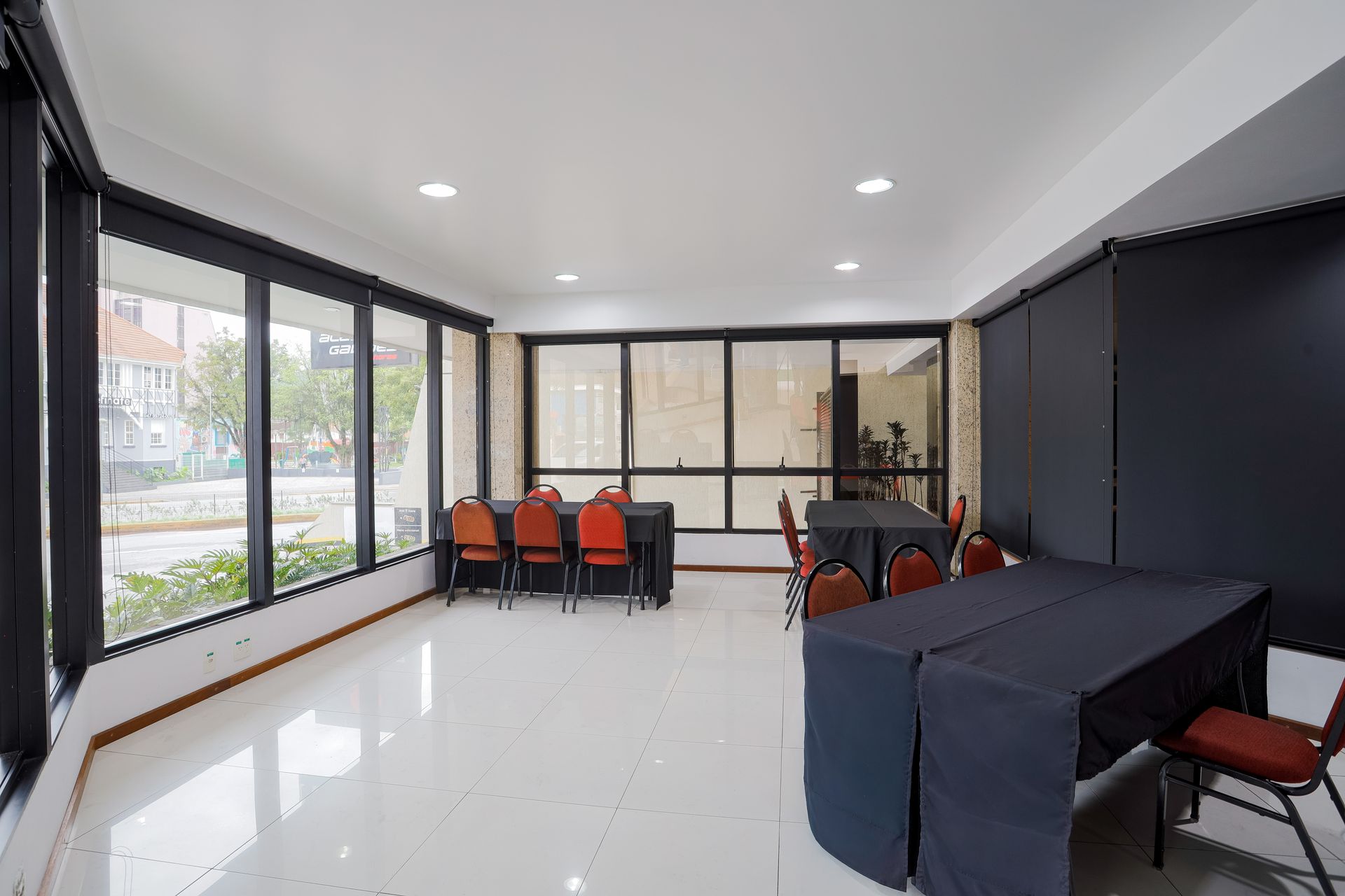 Quarto moderno e luminoso com piso de azulejo branco, paredes pretas, janelas amplas e cadeiras ao redor de mesas.