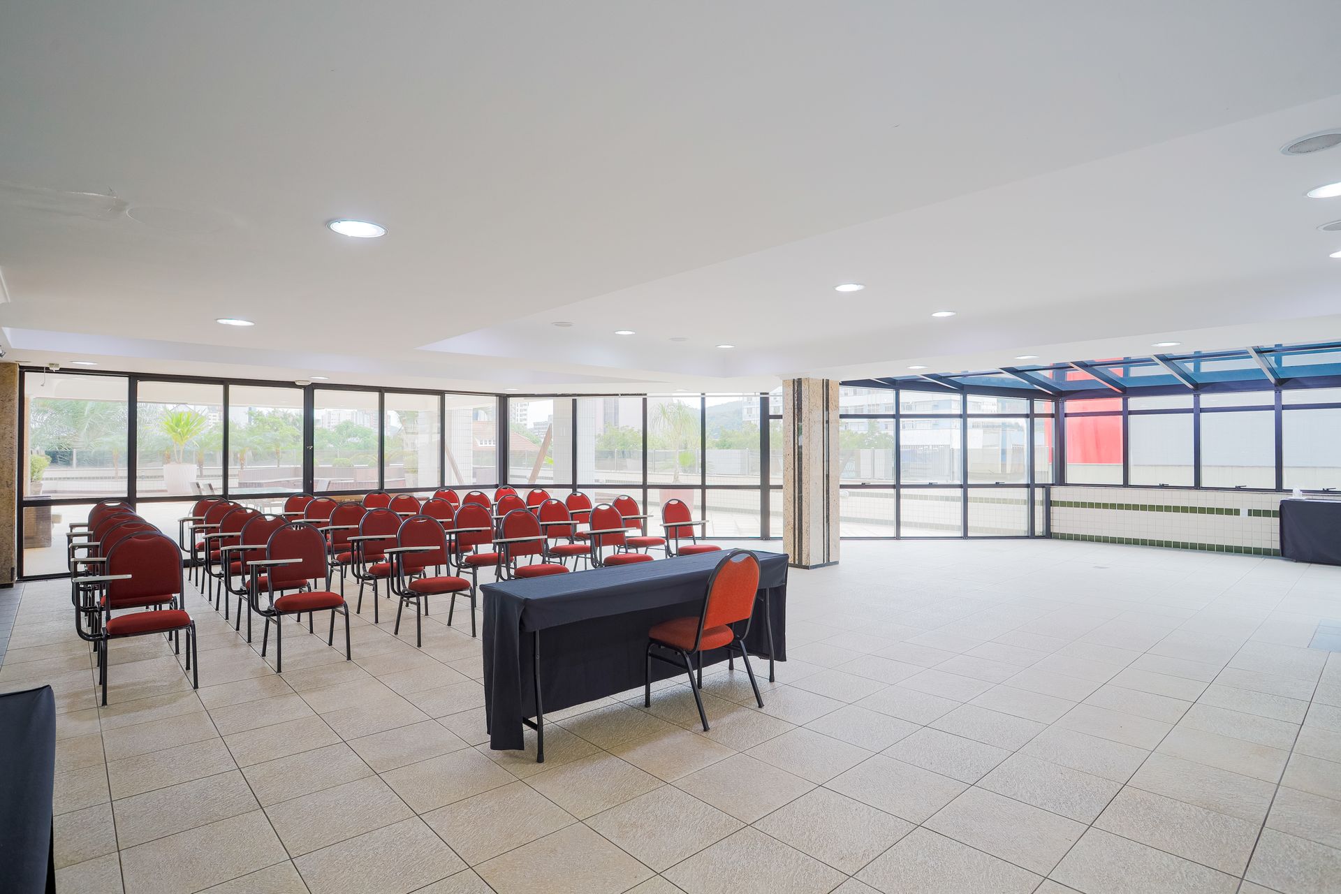 Salão de banquetes vazio com fileiras de cadeiras vermelhas e uma mesa coberta de preto perto de grandes janelas.