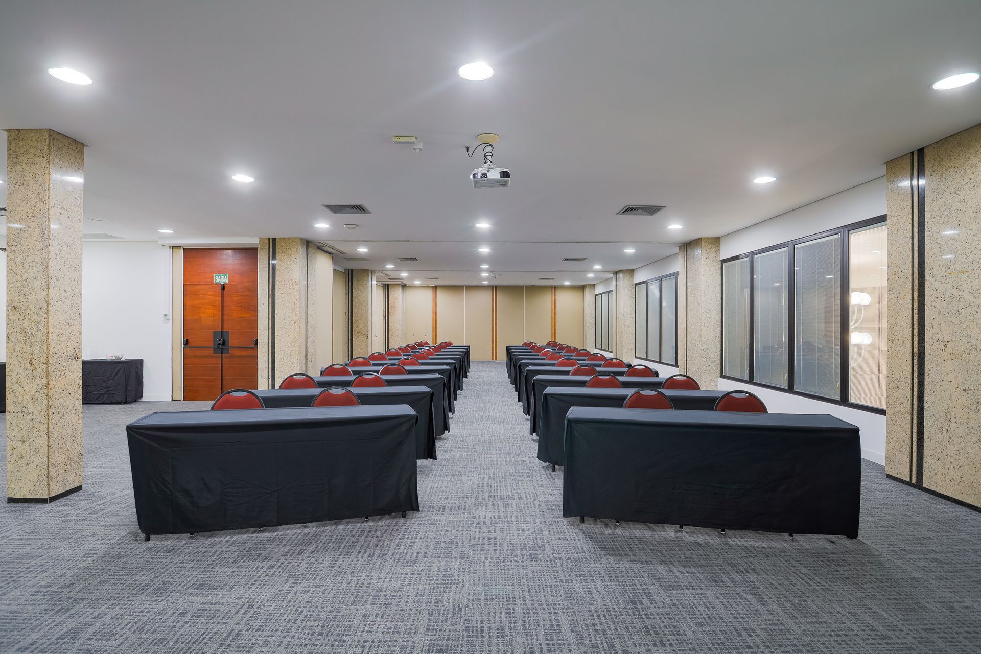 Sala de conferências vazia com fileiras de mesas pretas e cadeiras vermelhas sob luzes brilhantes no teto.