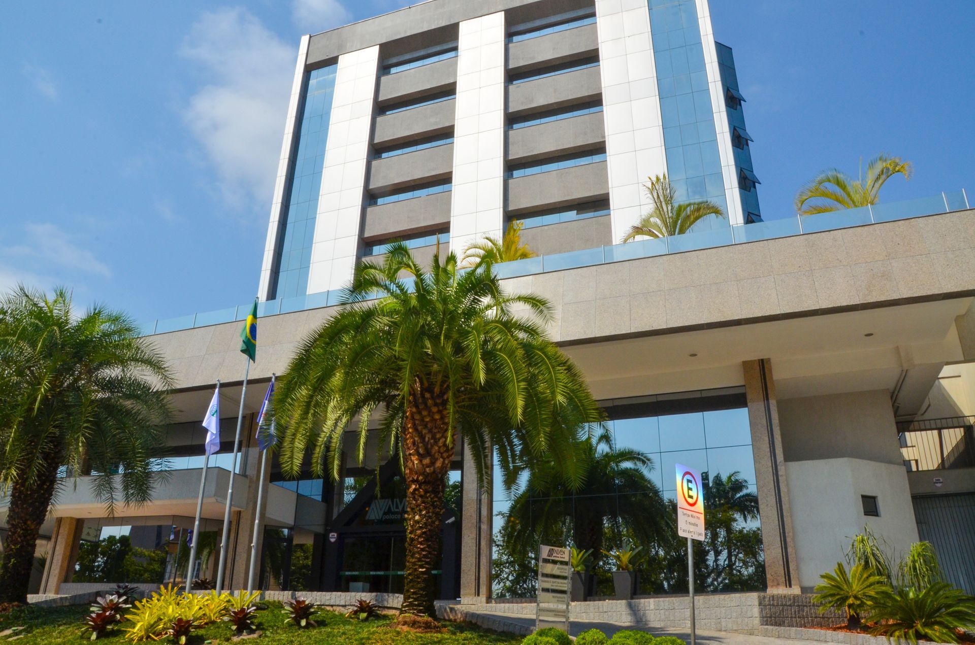 Torre moderna de hotel ou escritórios com palmeiras e entrada coberta sob um céu azul.