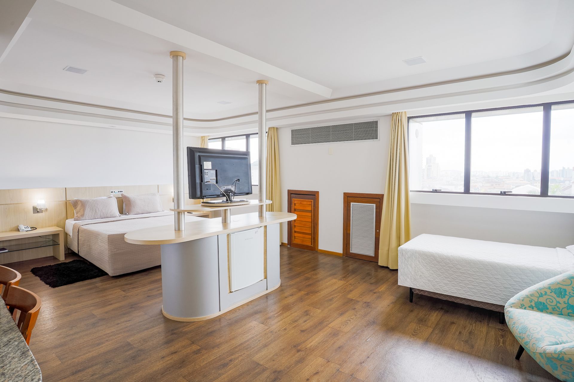 Quarto luminoso e mobiliado com piso de madeira, camas brancas, TV e janelas amplas.