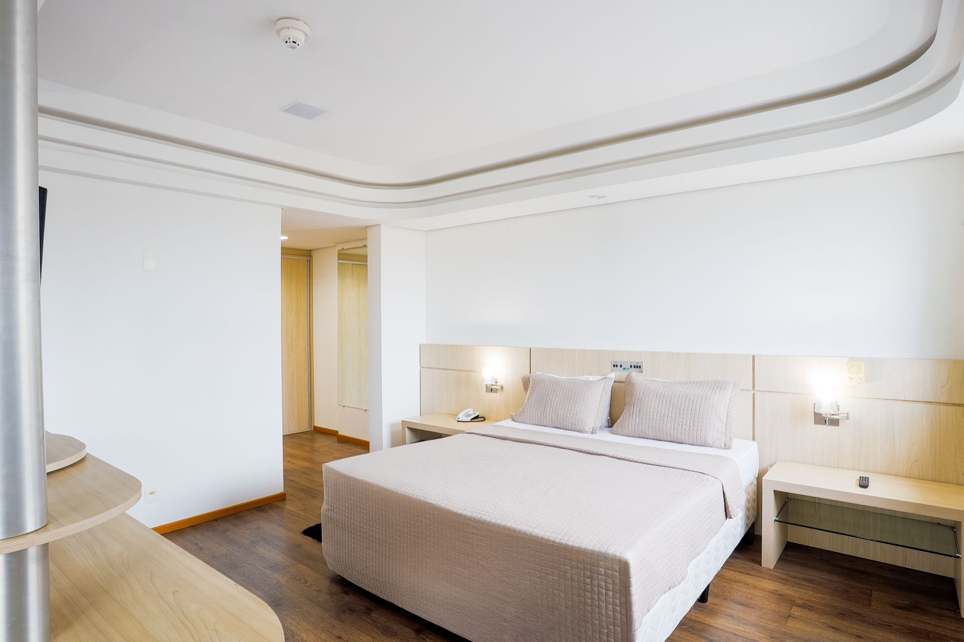 Quarto moderno e luminoso com uma cama grande, mesas de cabeceira e paredes brancas com piso de madeira.