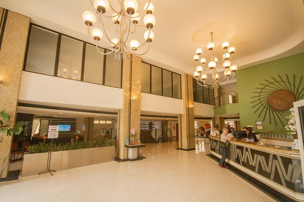 Hall de entrada luminoso do hotel com lustres, pisos de mármore, balcão de recepção e parede com detalhes em verde.