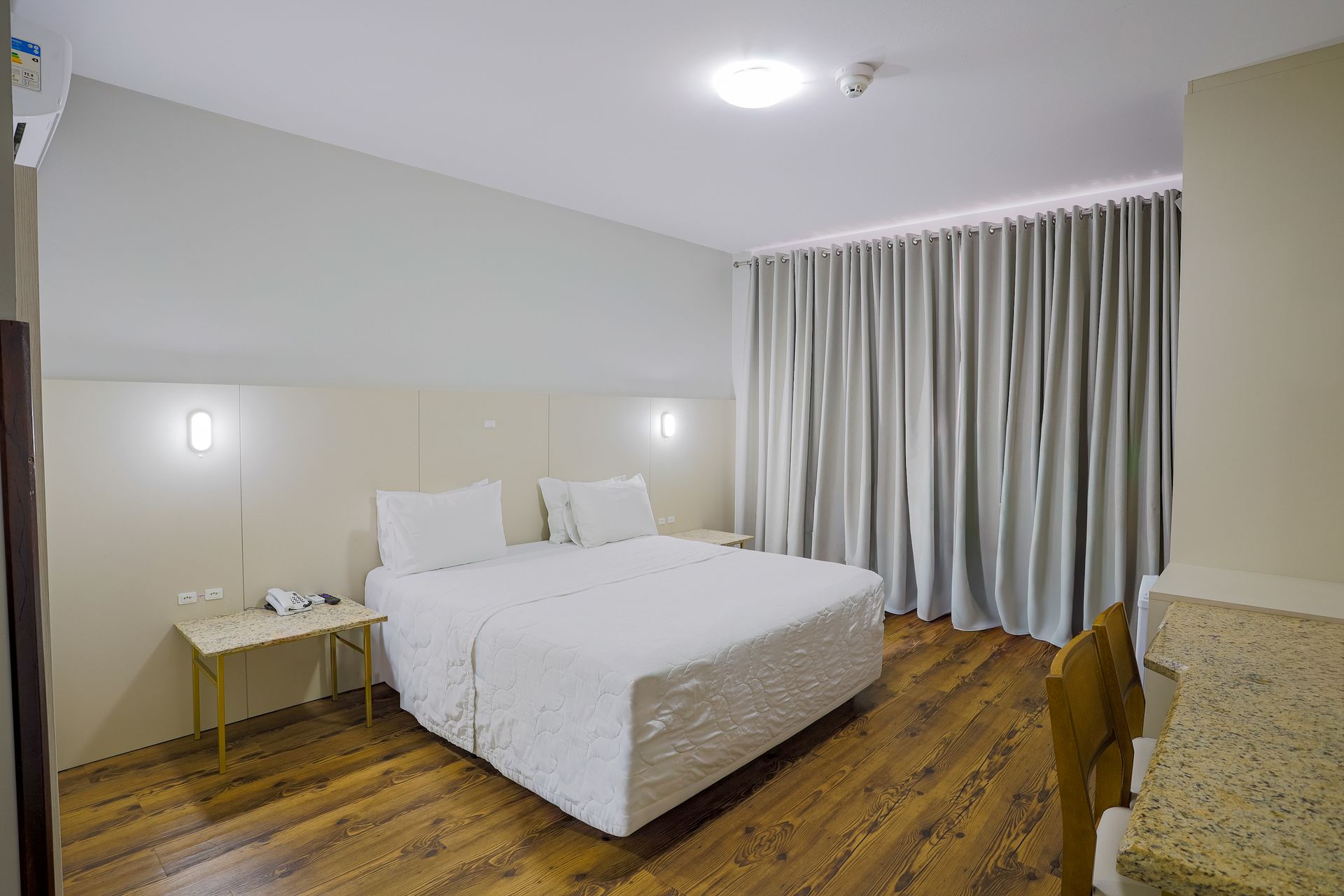 Quarto de hotel impecável com cama branca, mesa de cabeceira, cortinas e piso de madeira.