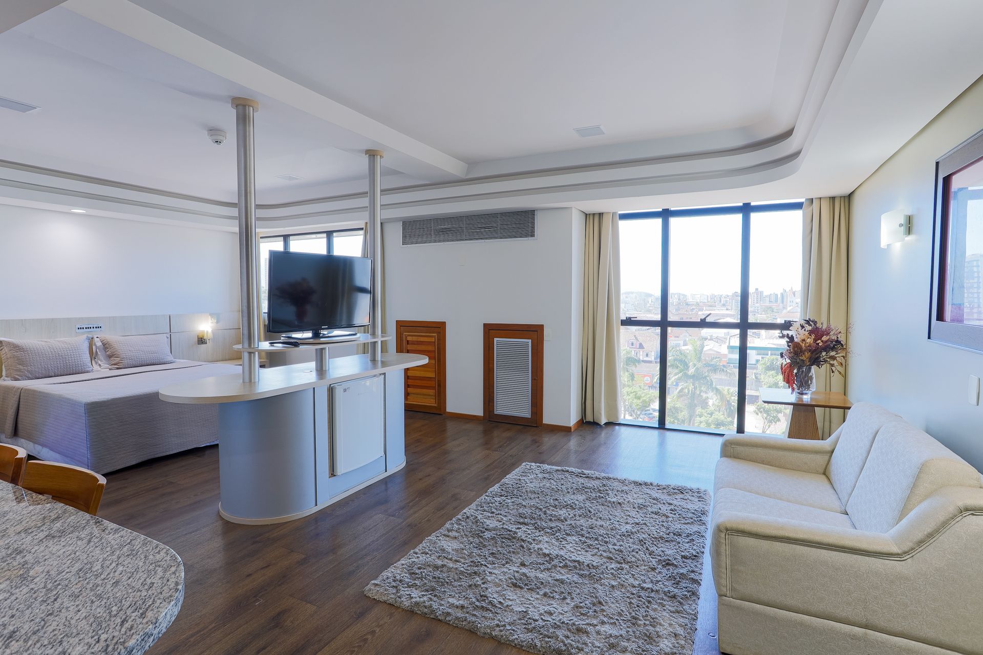 Quarto estúdio moderno e luminoso com sofá, cama, TV, tapete e portas de varanda com vista para a paisagem.