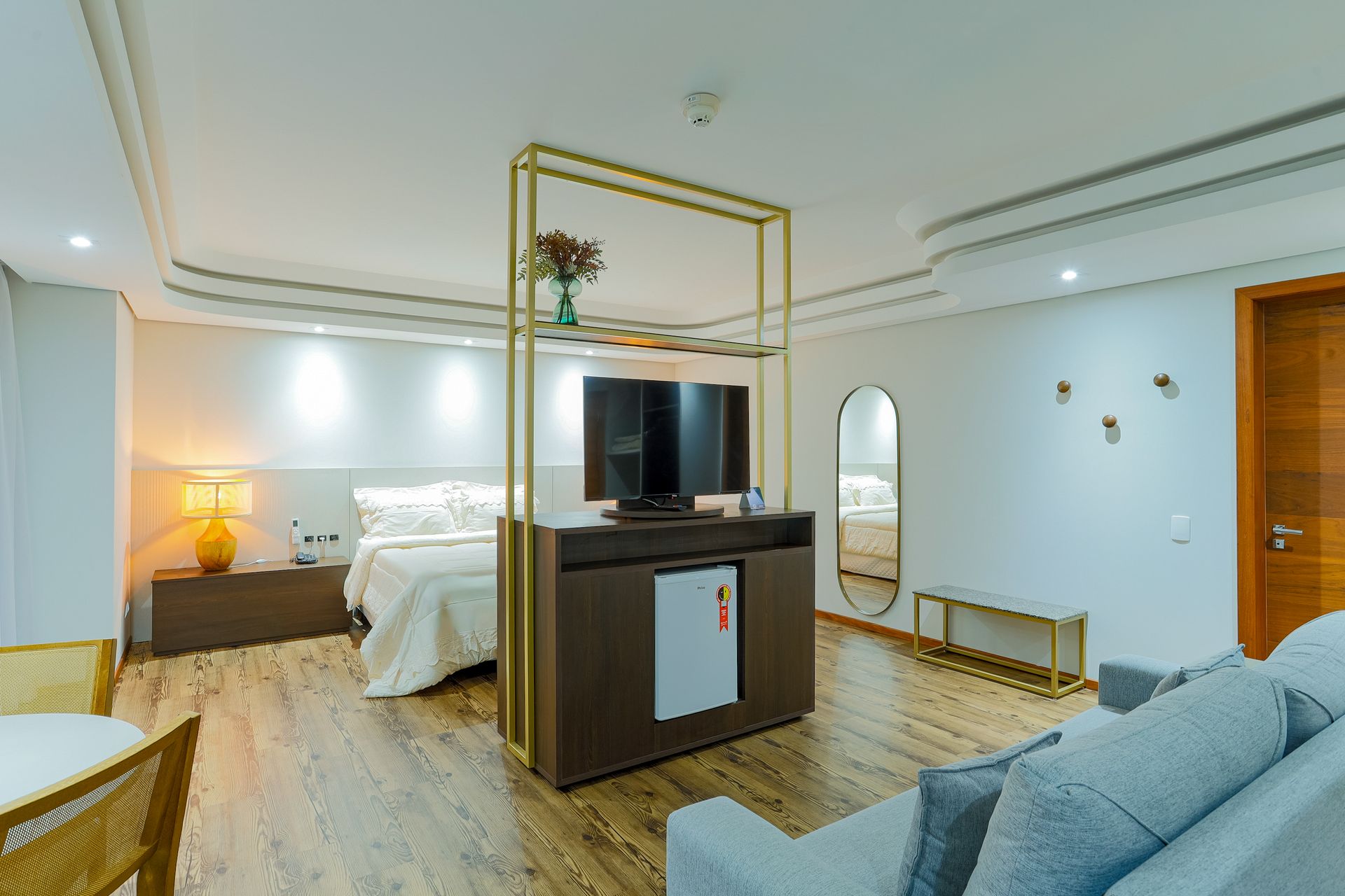 Quarto de hotel moderno com cama, TV sobre o armário, sofá e detalhes em madeira.